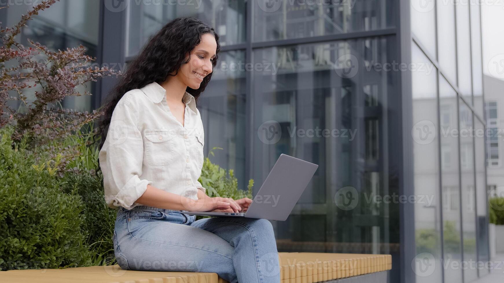 Smiling businesswoman sits on bench near office building on terrace with laptop chatting with clients colleagues works writes article. Girl student e-learning with wireless computer using free Wi-Fi photo