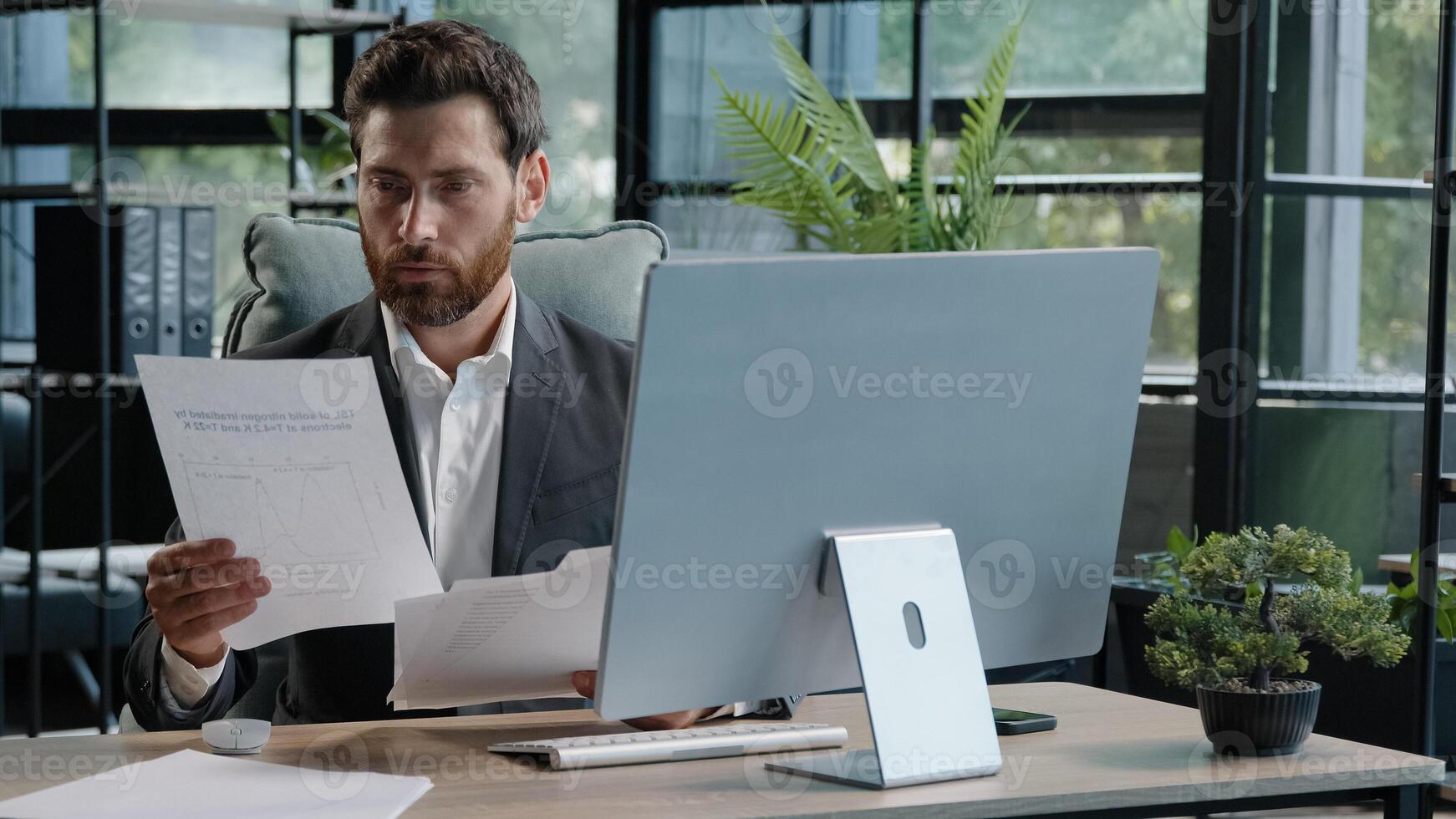 Serious adult 40s bearded pensive thoughtful businessman working at computer in office with paperwork. Professional employee worker typing thinking over online project check data in documents contract photo