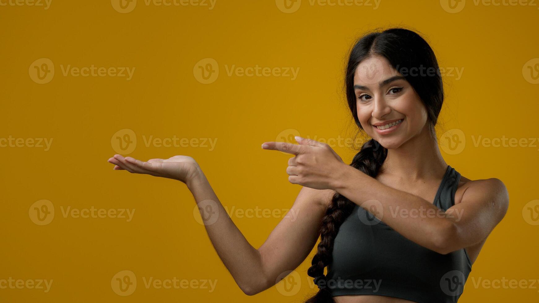 Portrait in studio Indian 20s woman posing on yellow background pointing to side at palm arm hand point aside recommend recommendation sign girl lady showing to empty copy space looking at camera photo