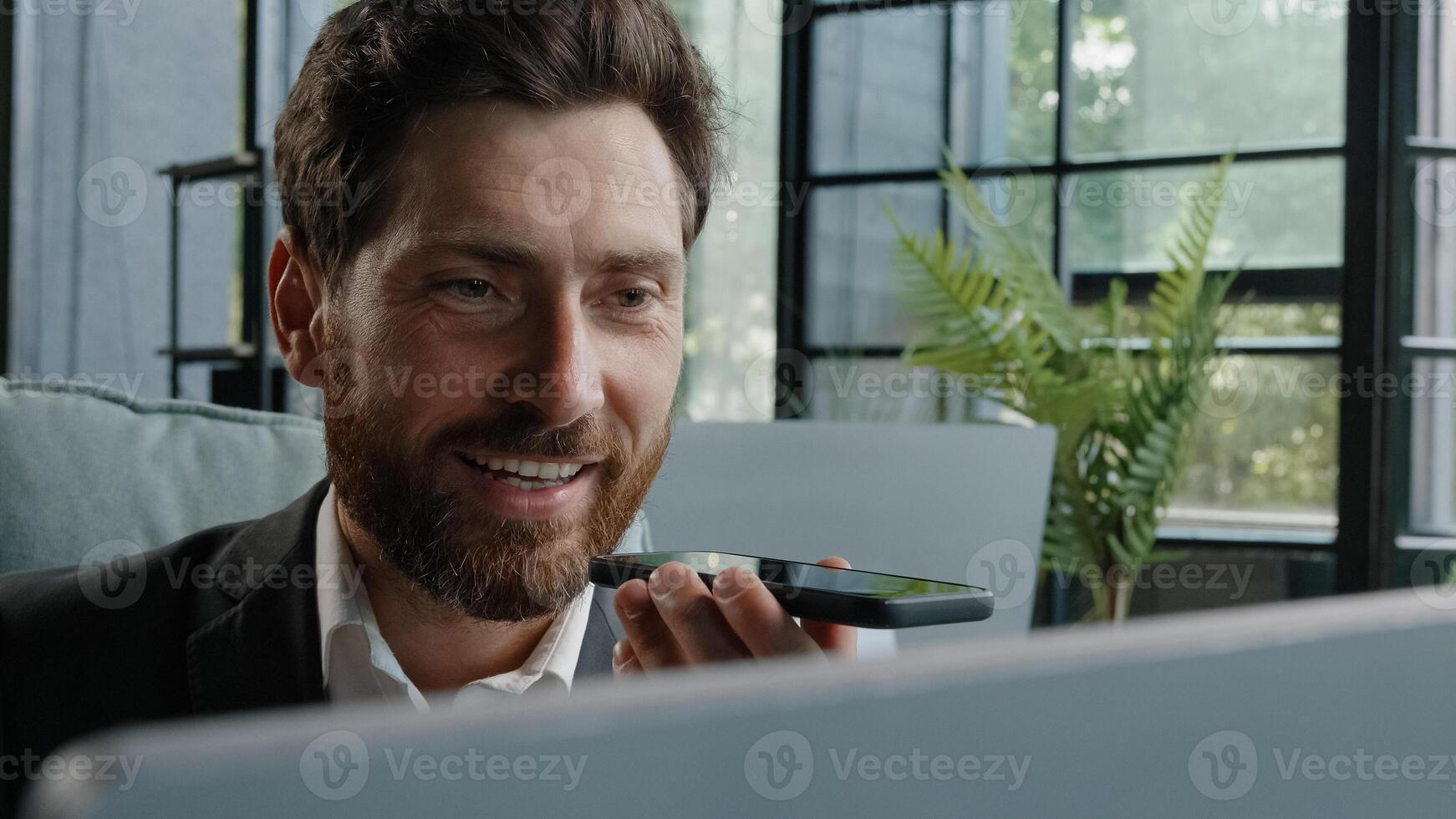 Happy adult 40s male manager bearded businessman sitting at workplace with computer smiling recording audio message sending voicemail using assistant software application on smartphone mobile app chat photo
