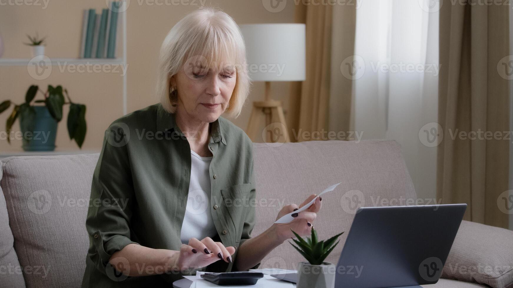 Caucasian old sad homeowner housewife makes woman budget checks monthly income by checking paper check using app laptop. Middle-aged businesswoman sitting on couch with document working remote at home photo