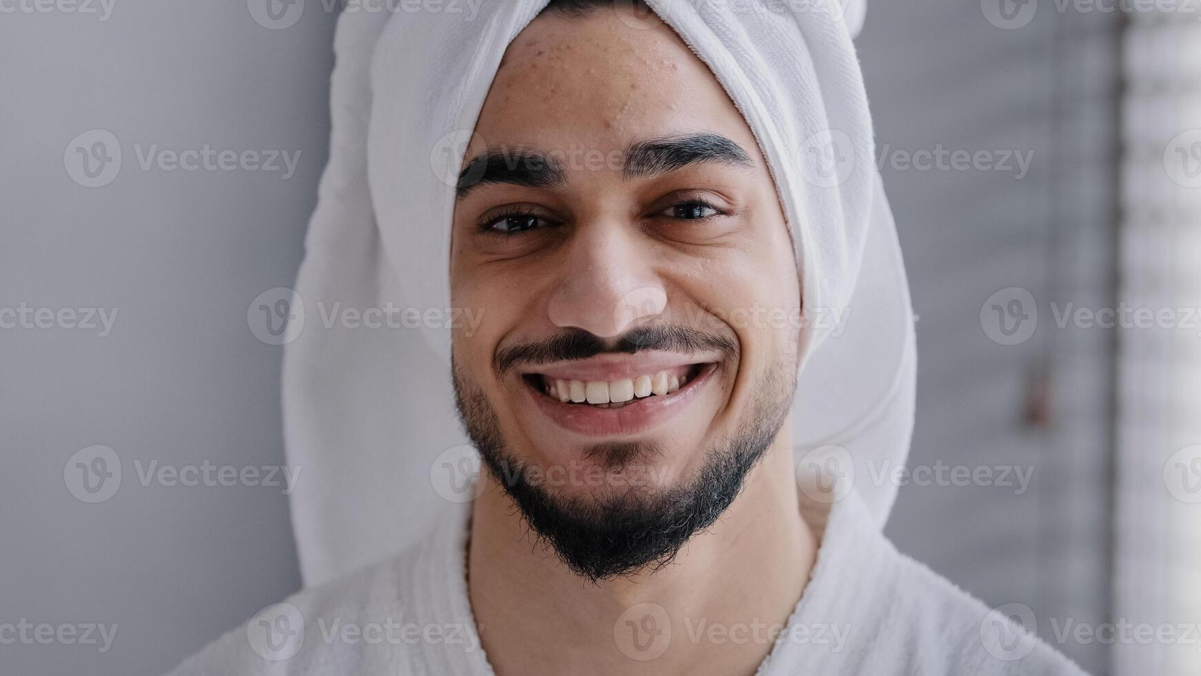 Closeup male face with acne headshot portrait smiling toothy handsome arabian indian bearded man with bath towel on head standing in bathroom. Healthy arab guy with white teeth smile looking at camera photo