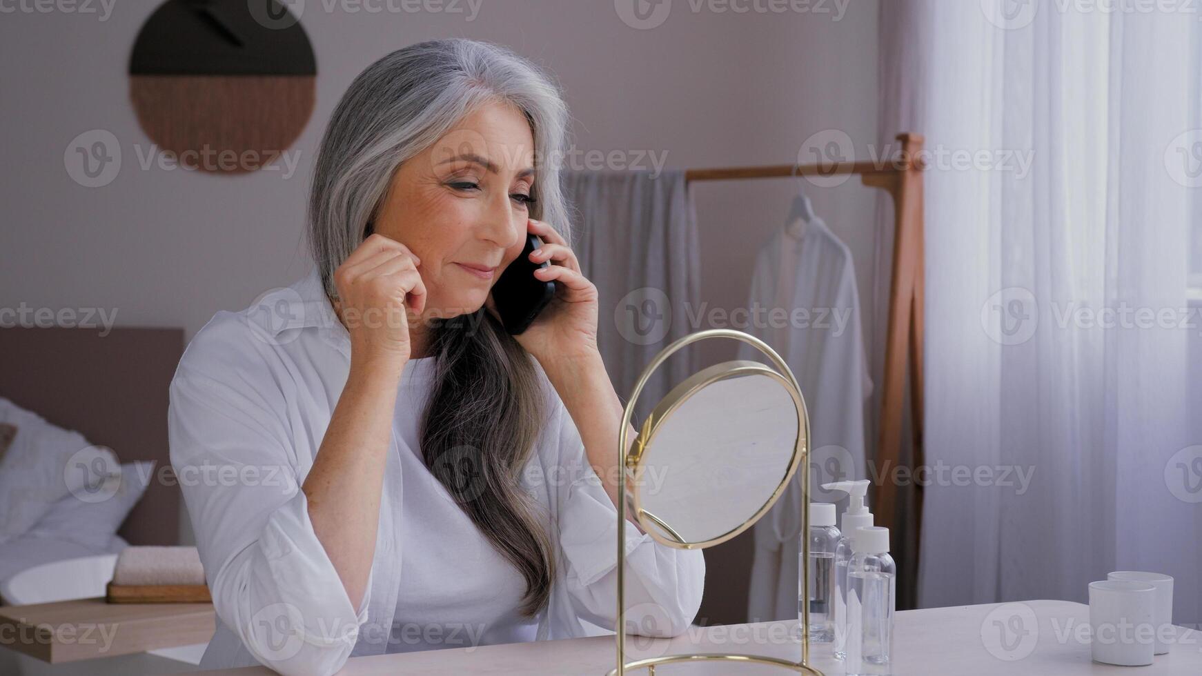 Senior gray-haired old 60s woman lady talking mobile phone talk conversation looking at mirror reflection buying ordering face cream by smartphone enjoy wrinkled beautiful aging perfect smooth skin photo