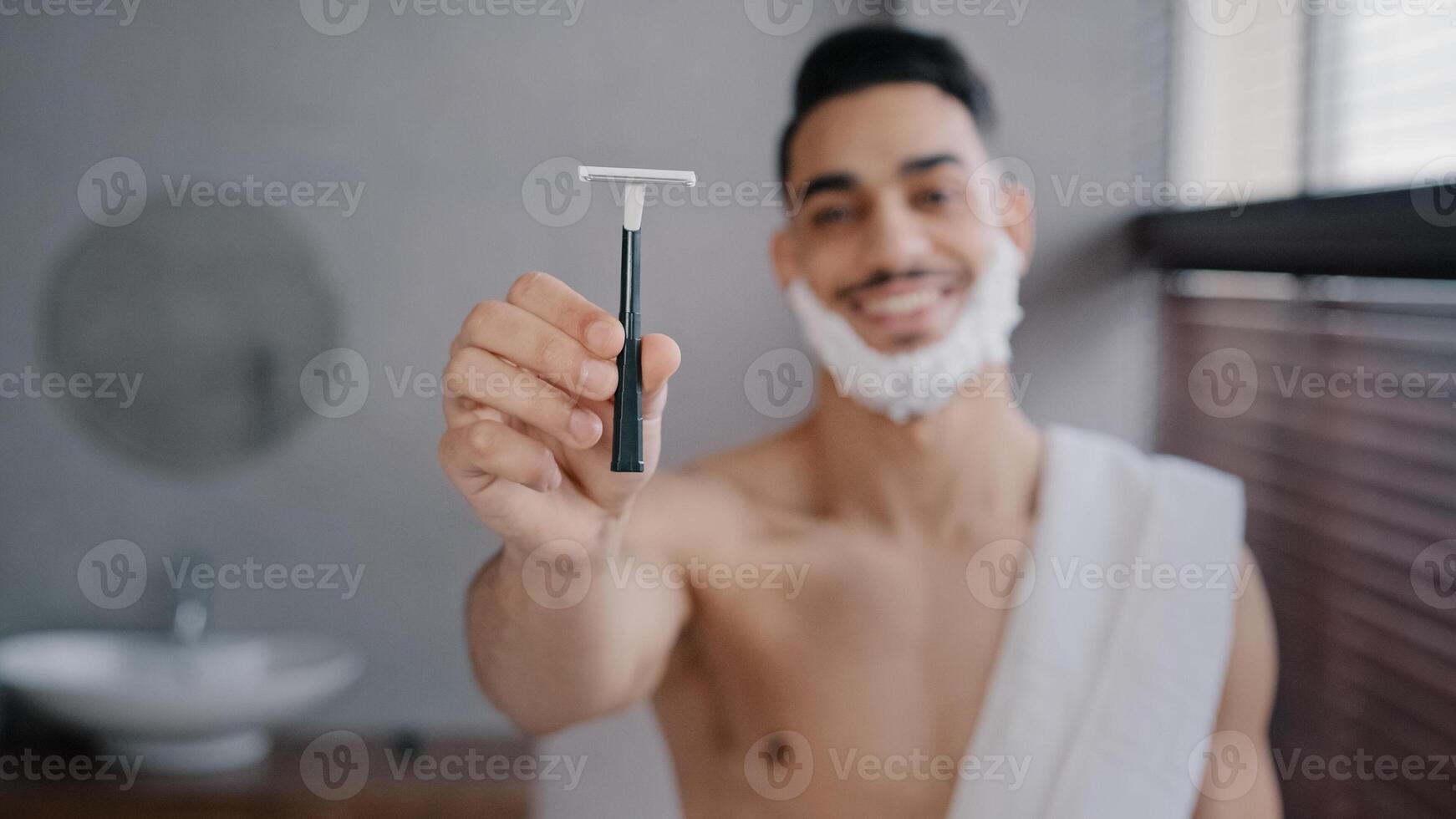 Funny hispanic indian arab naked man 30s bearded guy standing in bathroom at home with white soapy foam on beard shaving gel smiling showing razor looking at camera getting ready to shave facial care photo
