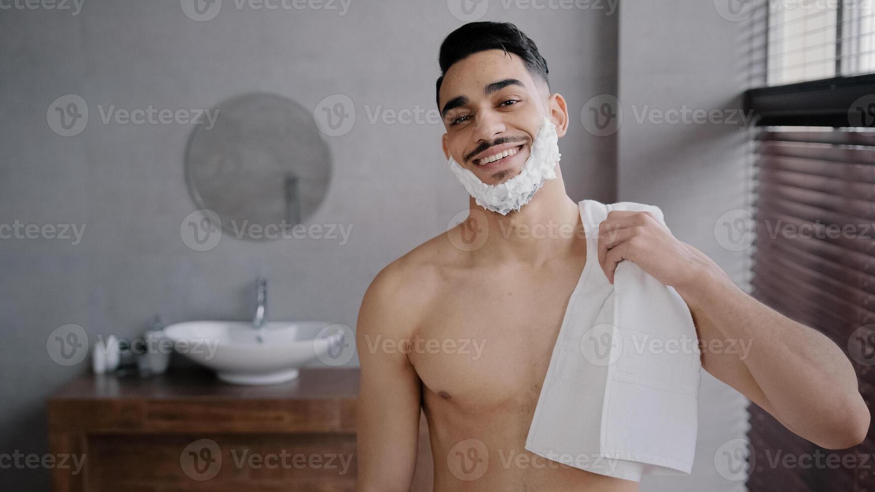 Close up male portrait in bathroom hispanic indian arabian naked man bearded guy posing smiling with white soapy foam on beard shaving gel holding bath towel looking at camera getting ready to shave photo