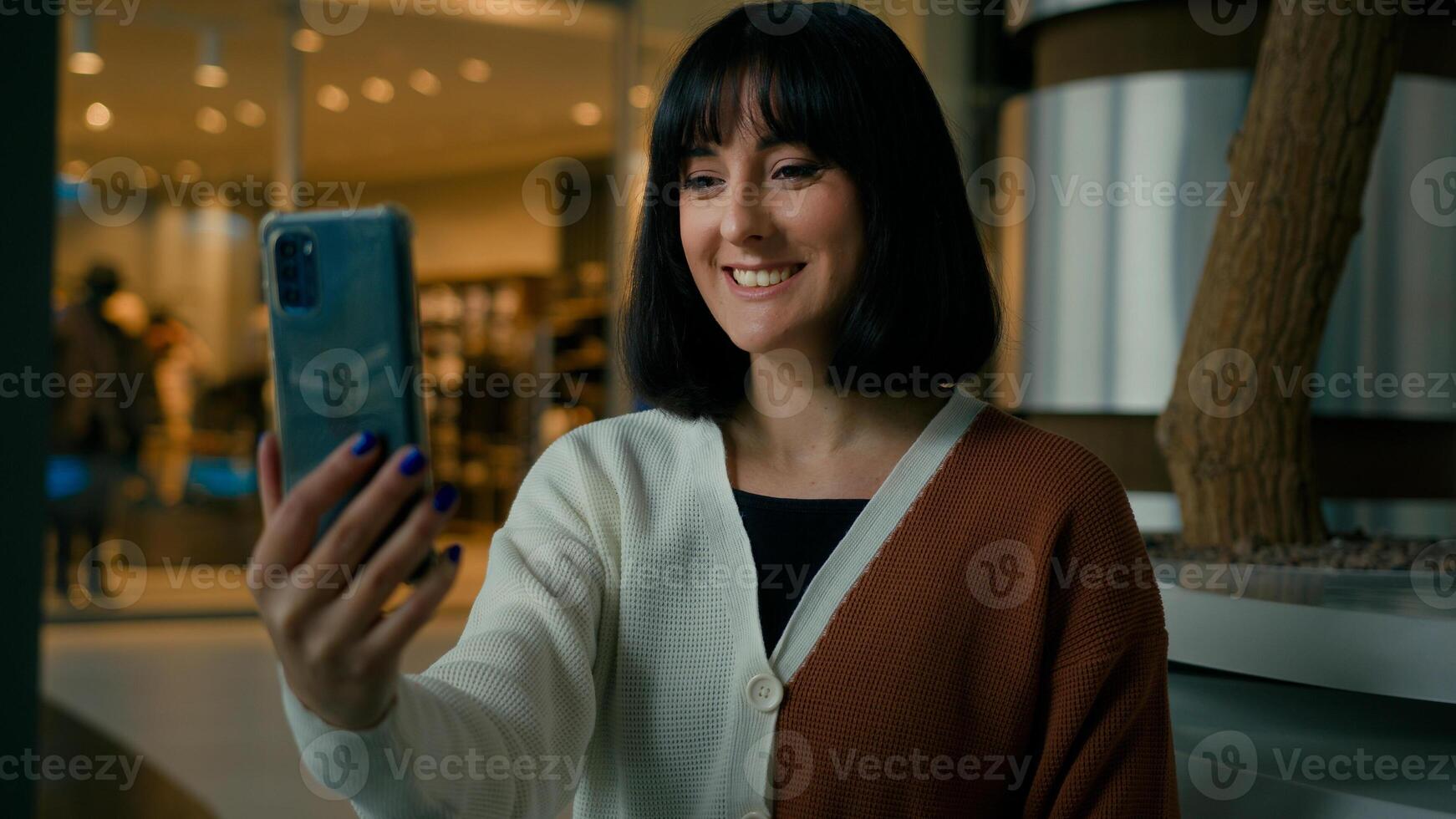 Smiling positive talking to smartphone webcam woman has call indoors in shopping mall waving hand hello hi greeting girl speak phone conference distant chat virtual meeting talk remote speaking photo