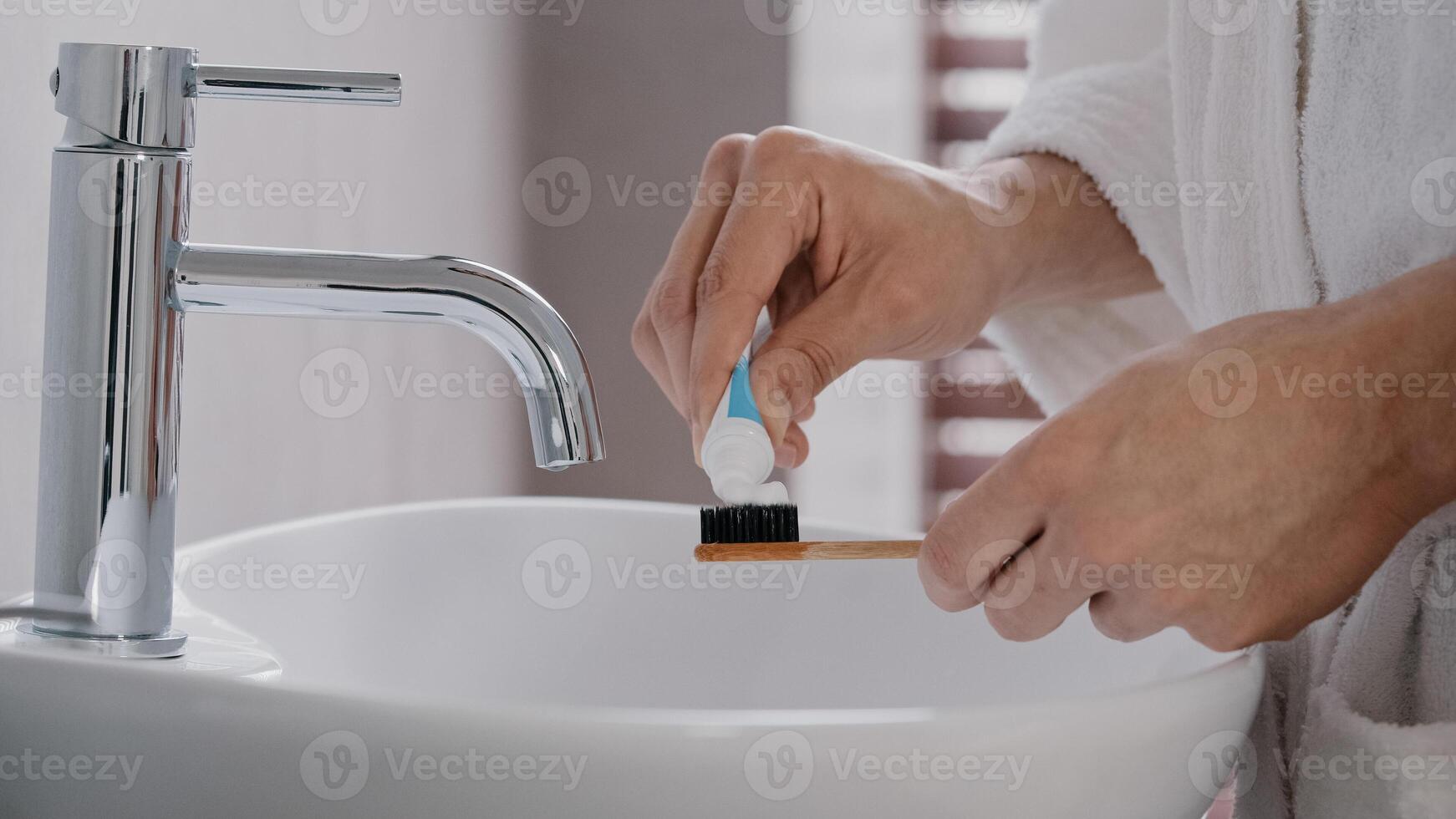 Close-up of male hands in bathrobe holding bamboo wooden dental tally squeezing out tube of whitening toothpaste preparation for brushing teeth in morning near sink in home bathroom tooth dental care photo