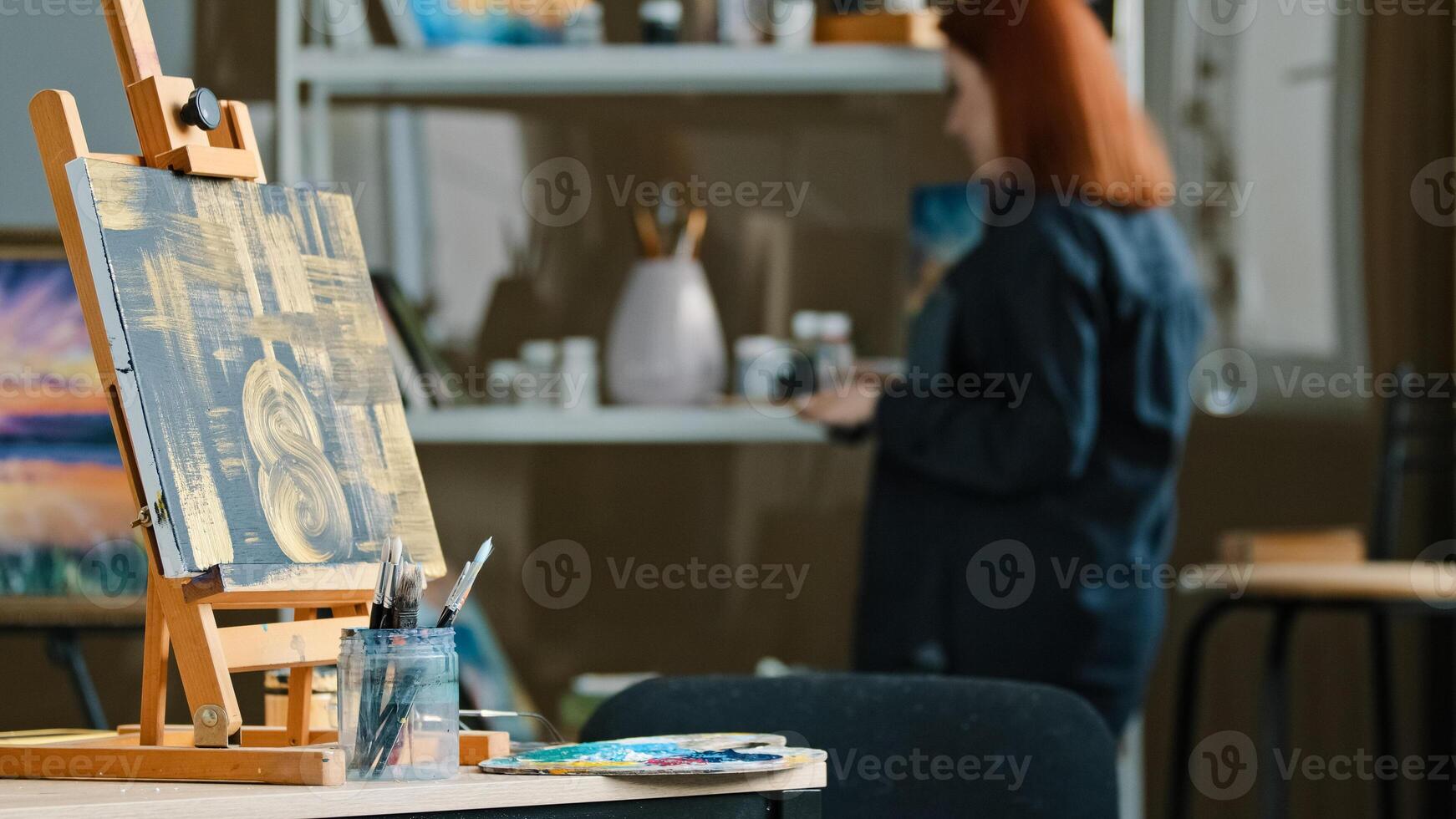 Abstract painting black canvas with gold drawings on easel stands on table in workshop studio with paint brushes red-haired unrecognizable woman artist painter choose acrylic oil preparing for drawing photo