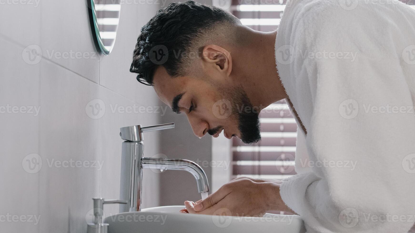 Side view arab spaniard indian arab man guy opens faucet near washbasin at home in hotel bathroom washes face with hot cold water morning beauty ritual male hygiene guy in bathrobe wet facial wash photo