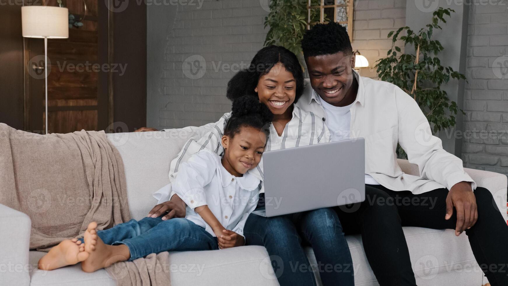 Happy African family and cute kid child girl daughter using laptop looking at computer screen enjoying watching funny social media having online call conference relaxing on sofa at home photo