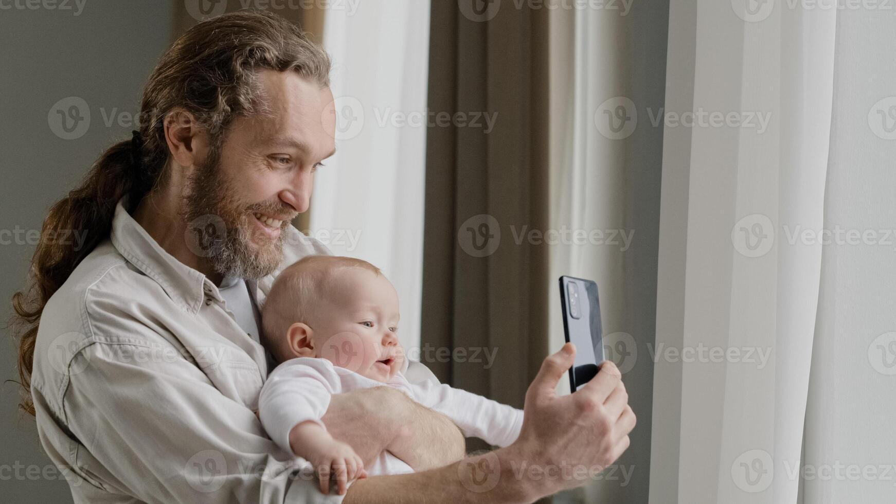 Happy smiling caucasian father adult daddy holding newborn child at home standing near window dad with little daughter son infant newborn making photo vlog online stream conference distant call