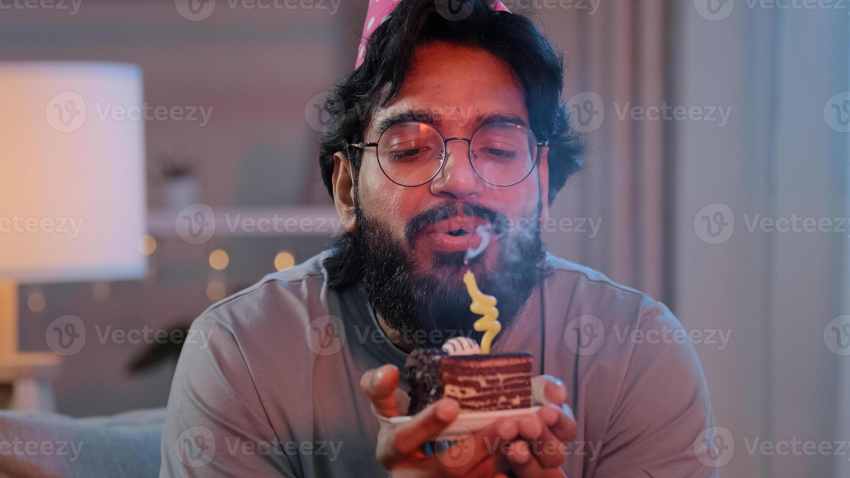 Portrait arabian happy indian male face bearded man wears festive birthday pink hat and glasses holds cupcake makes wish joyful congratulating at home blowing burning candles on cake celebrating party photo