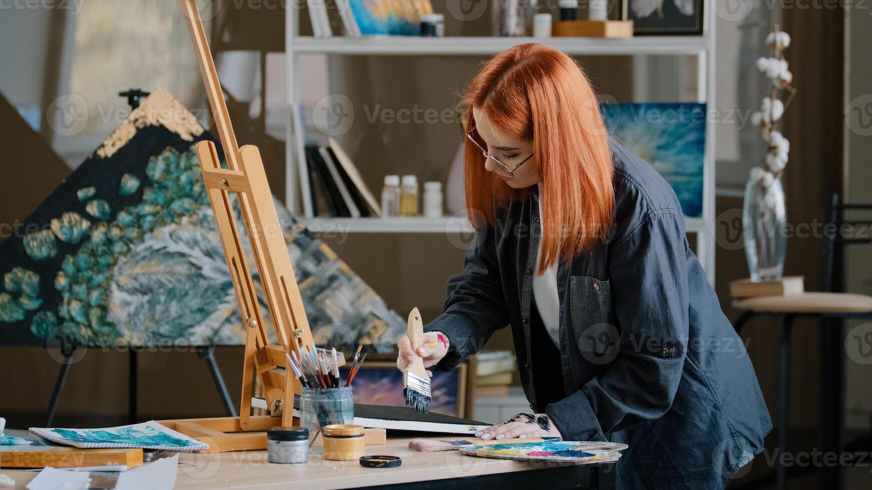 Inspired busy focused redhead girl artist woman painter in art studio background paints with wide brush on canvas with acrylic oil colorful paint puts picture on easel satisfied looking at camera photo