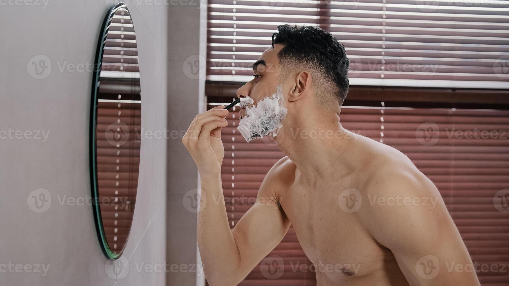 Close up side view bare hispanic guy face skin care arabian arab bearded man shaving beard using disposable razor in bathroom looking at mirror at morning routine. Indian male hygienic beauty shave photo
