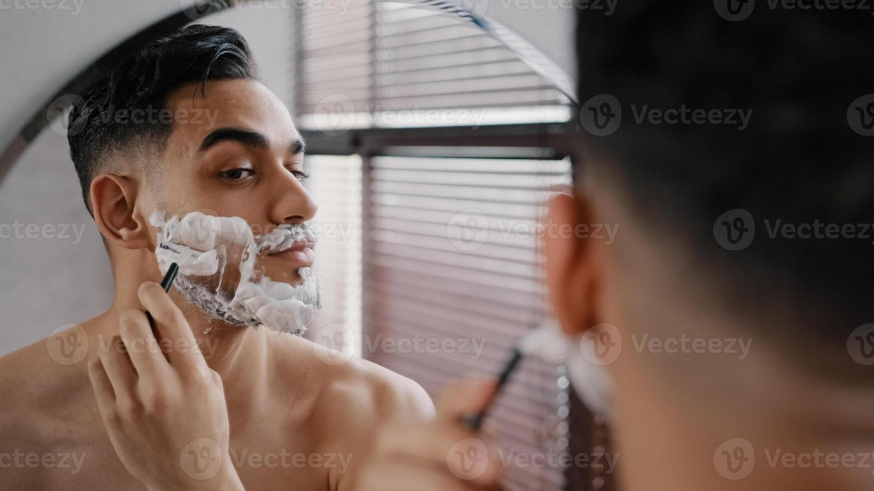 Hispanic arabian arab indian bearded handsome man shaving using disposable razor in bathroom looking at mirror. Daily morning routine male hygienic beauty procedure. Guy shaves beard and moustache photo
