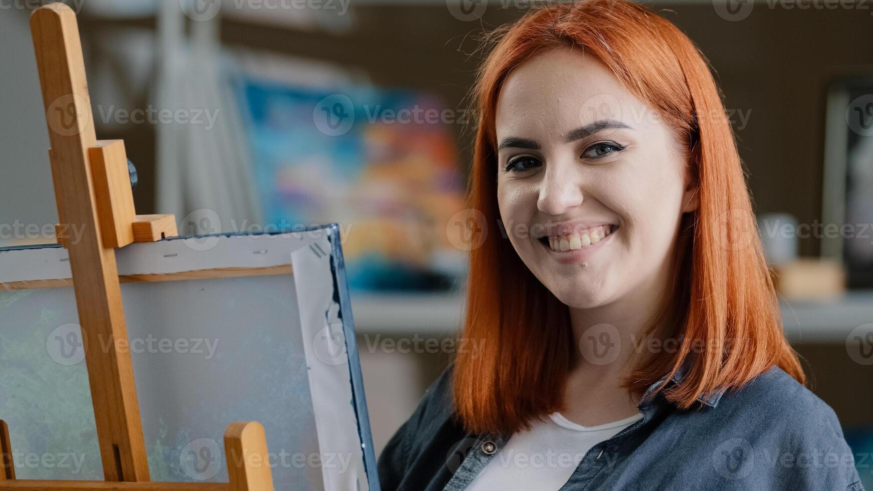 Caucasian woman artist girl painter with red hair enjoying art work with paints creates picture drawing painting on canvas looking at camera smiling posing smile toothy happy cheerful draw laughing photo
