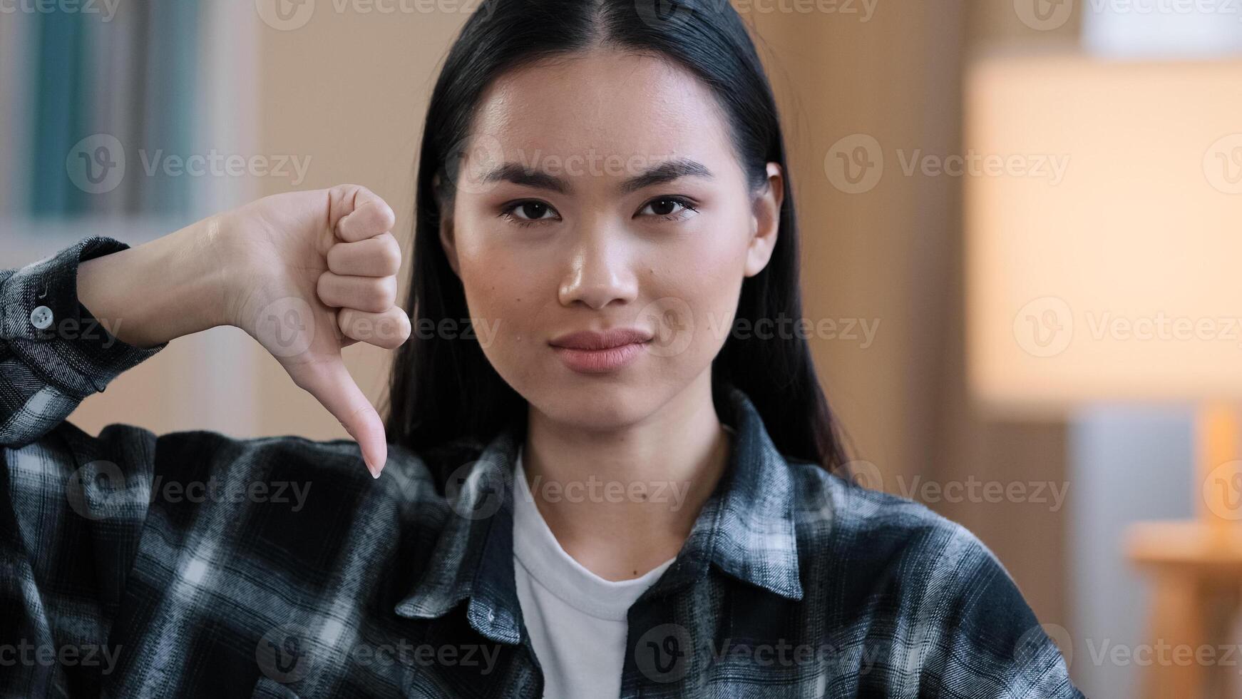 Close up asian millennial woman korean girl wear casual plaid shirt looking at camera showing dislike thumb down disagreement disgust rejection sign bad attitude gesture refusal failure symbol indoors photo