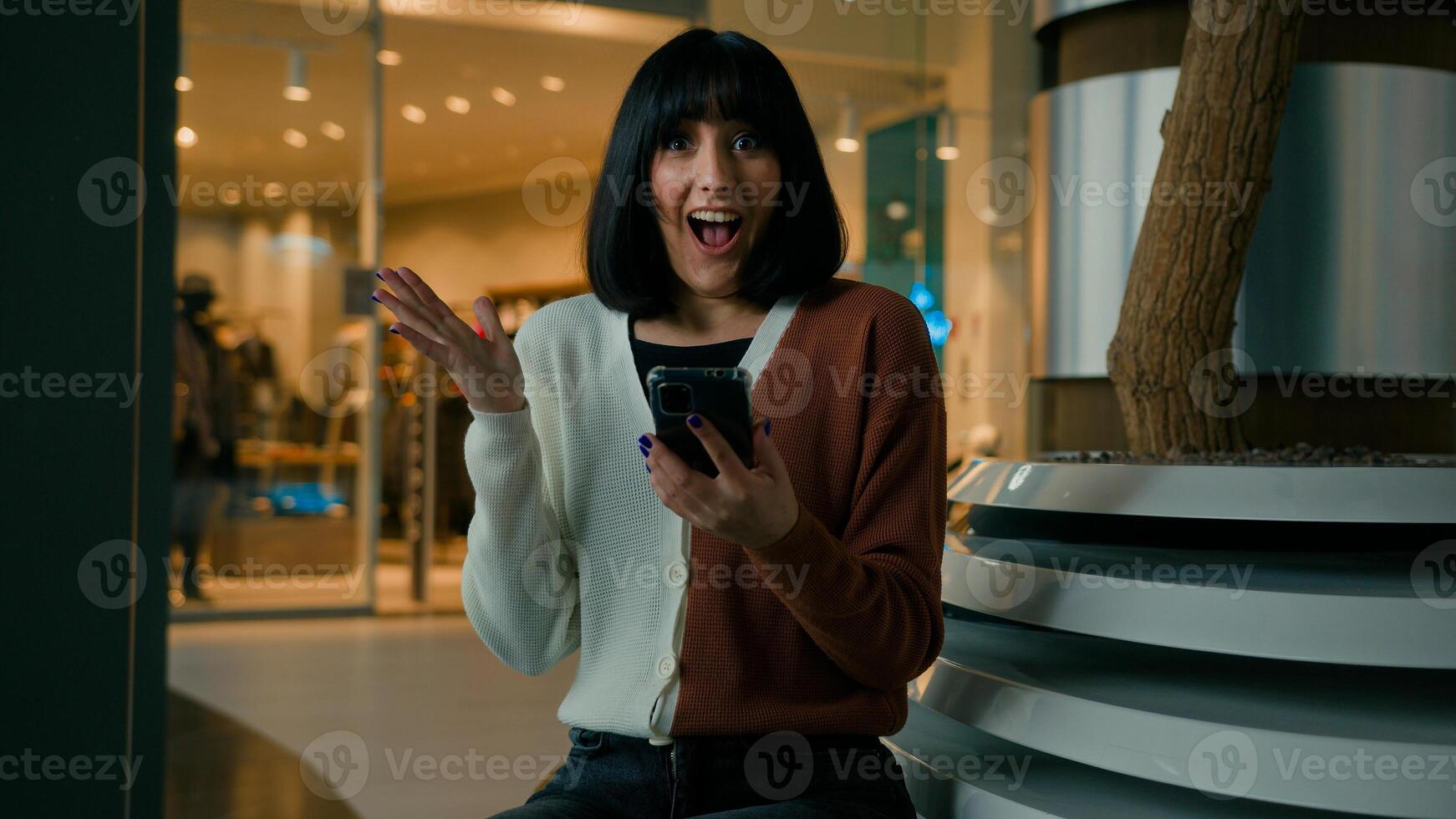 Woman indoors with phone shocked reaction female girl win with mobile phone bet winning with cellphone wonder wow with smartphone good news looking at camera with shock pointing finger on cell screen photo