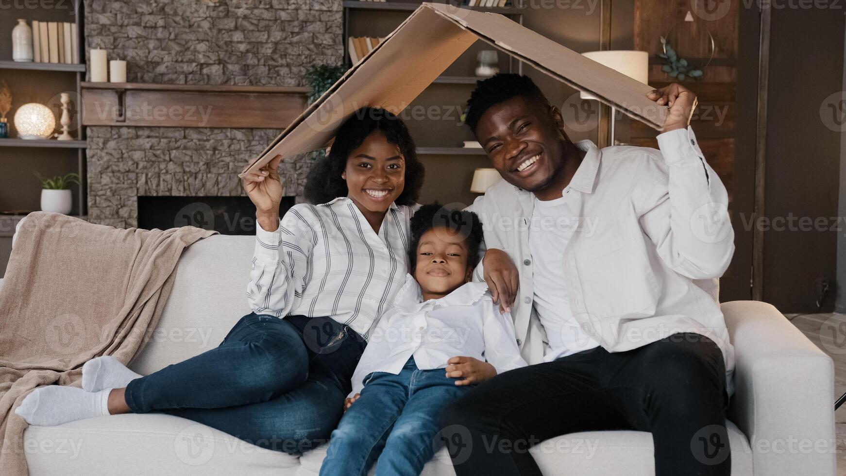 African couple sitting on couch under cardboard roof smiling looking at camera active happy daughter girl running sits on sofa. Relocation day to new home moving to own apartment homeowner portrait photo
