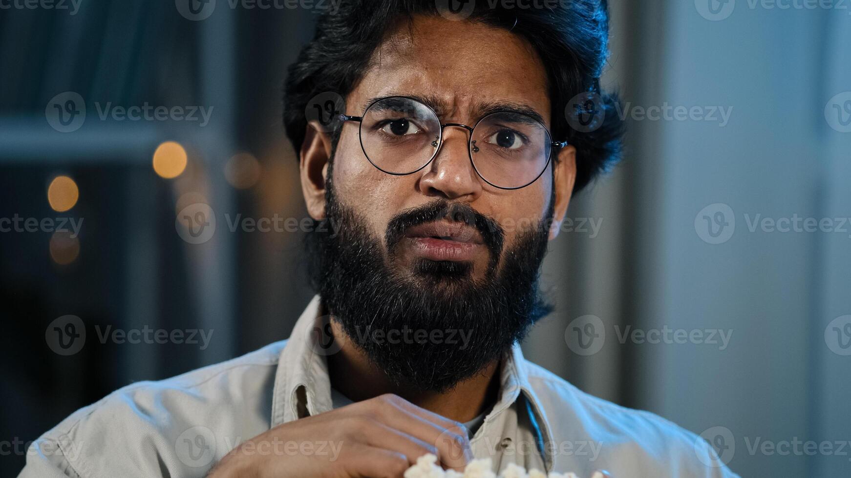 Male home portrait serious interested attentive Arabic Hispanic Indian bearded man guy in glasses eating popcorn watching TV show series late evening night watch movie online sport game championship photo