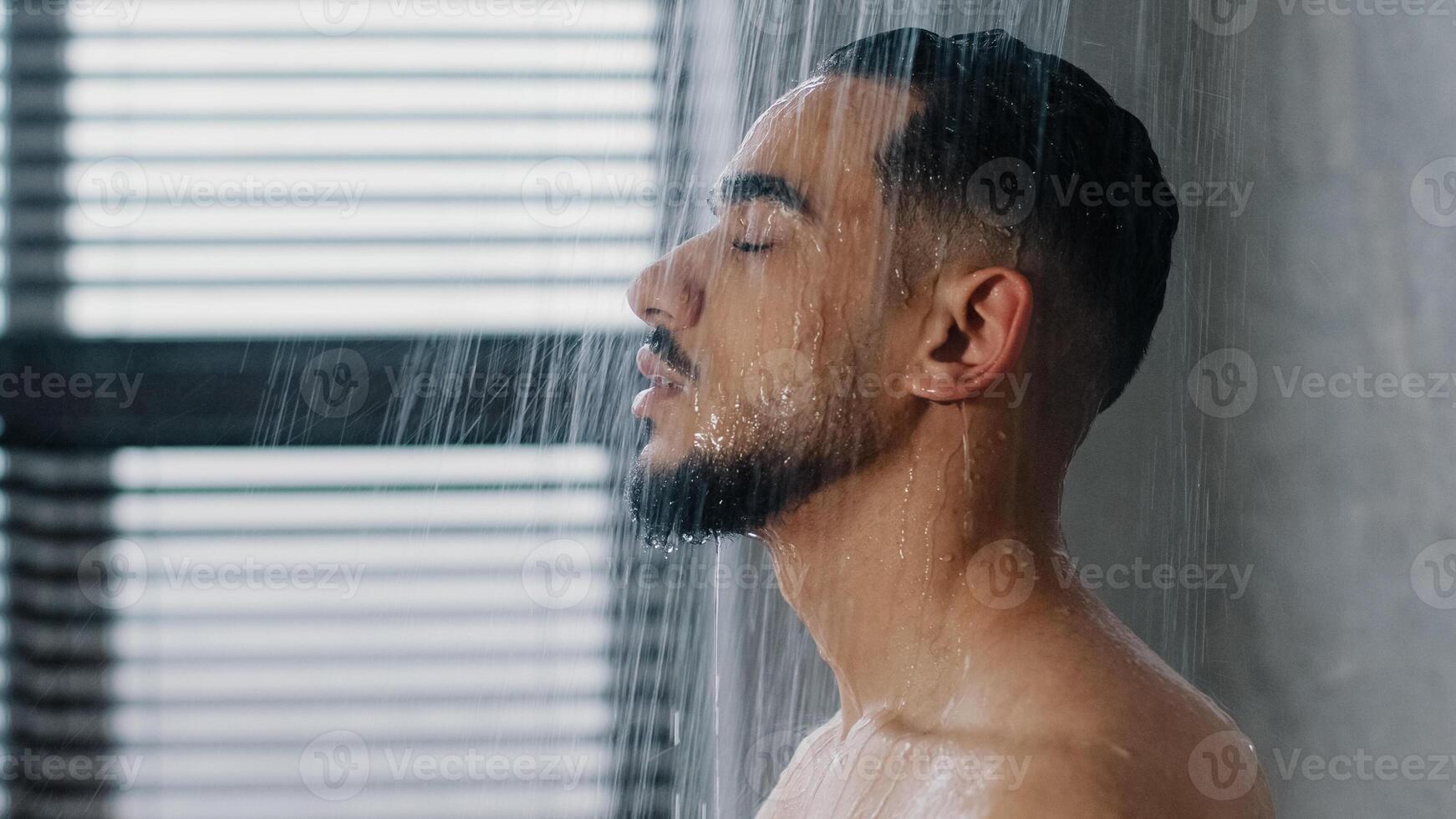 Side view indian relaxed man bearded guy naked male washes head hair face and beard in bathroom showering in at home in hotel after workout close up. Brunette rinsing shampoo and conditioner photo