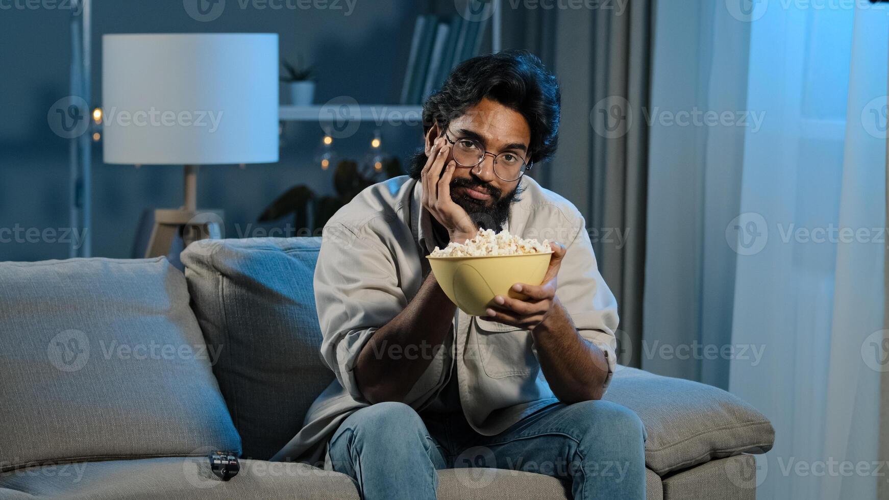 Upset sad bored Arab man tired indian male with beard sits at home evening watching boring TV program switches channel laughing enjoying comedy movie eating popcorn relaxing in dark living room alone photo