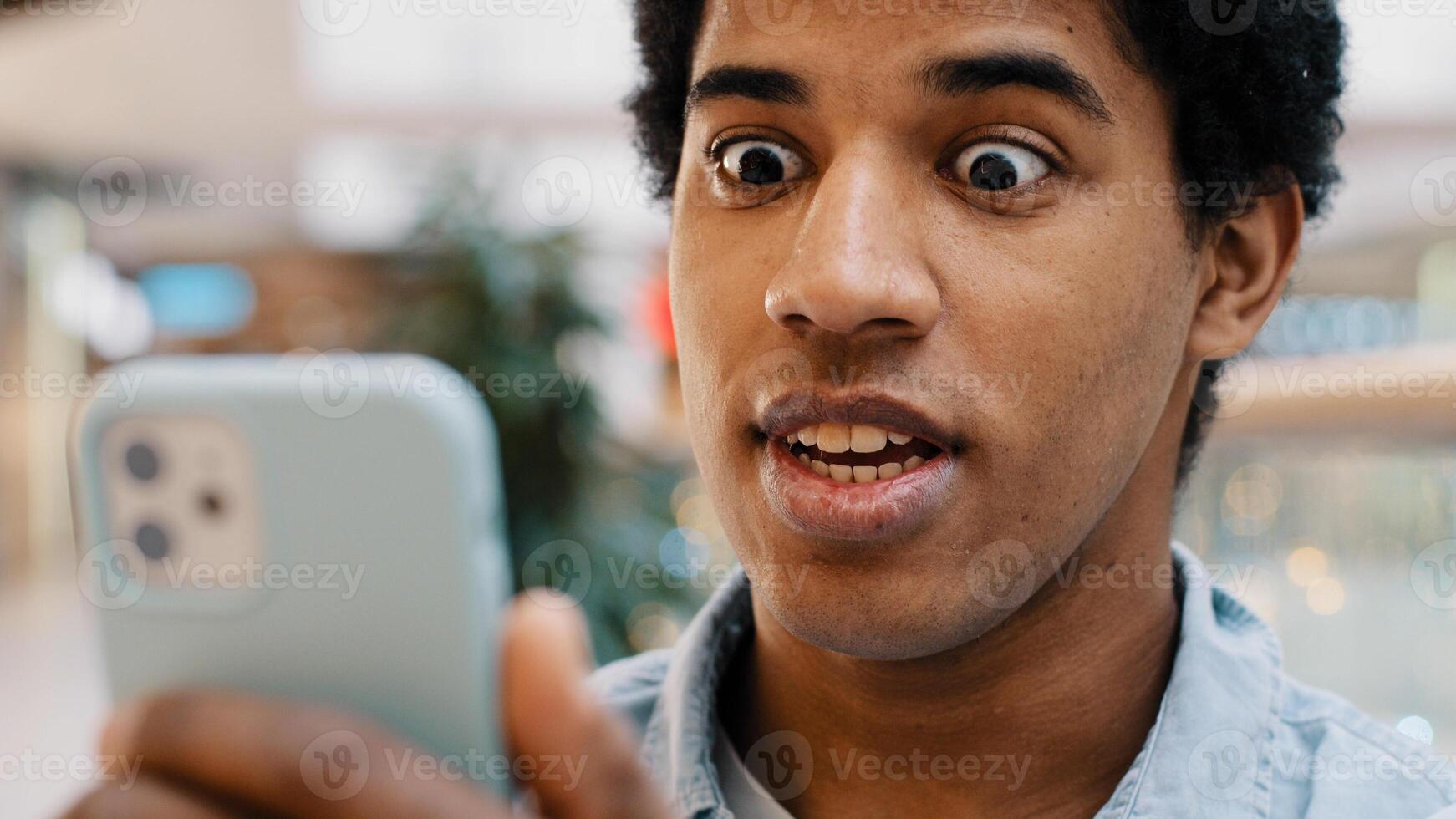 African happy excited guy surprised by message good news winning online bets looking at mobile phone has offer invite opens mouth in surprise delighted shock man feels amazing wonder with achievement photo