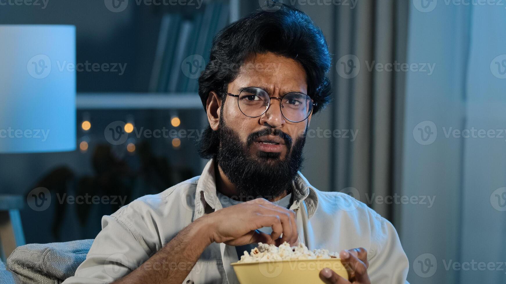 Interested attentive Arabic Hispanic Indian bearded man guy in glasses eating popcorn watching TV show series horror movie late night feeling scary shocked scared reaction disgust horrible thriller photo