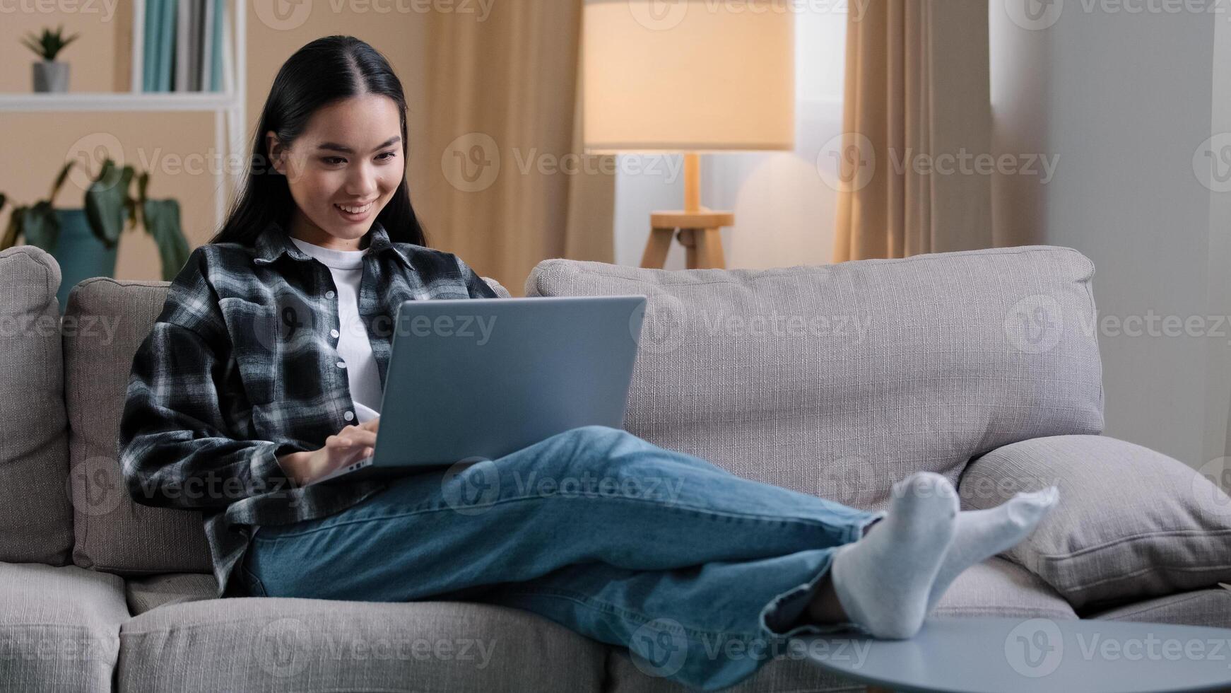 Carefree Asian girl enters living room sit down on comfortable sofa takes laptop with smile and watches online woman uses computer sitting on couch at home casual browsing net resting typing photo