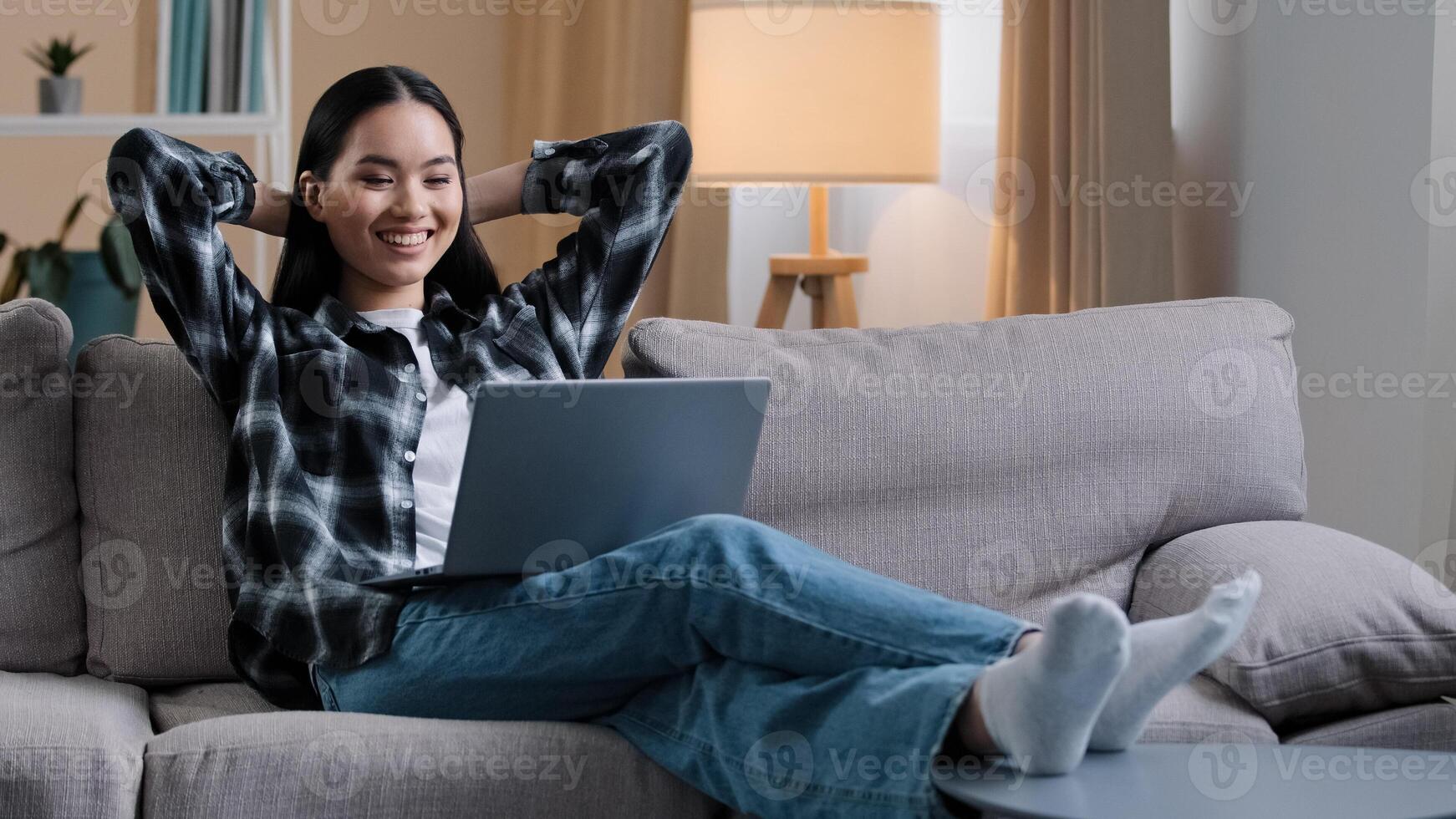 Asian woman Korean girl freelance student lady user sitting on sofa with hands behind head resting work break stop e-learning watching movie online laughing enjoying leisure time with computer photo