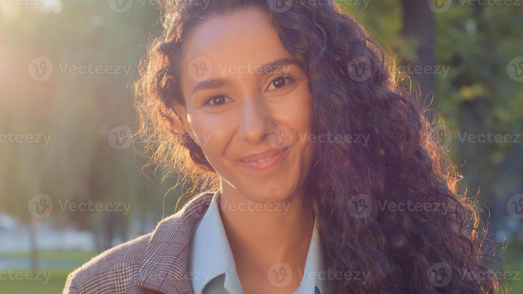 Beautiful female lady girl looks at camera with smile posing in sun lightssmiling outdoors. Woman with long black curly hair in sunbeams in park posing happy. Attractive model with flying curls photo