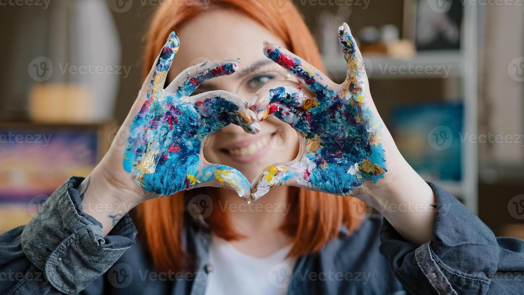 Close up head shot red-haired 20s smiling girl in eyeglasses smiling toothy friendly with love painter woman looking at camera holding dirty in paints hands in heart shape shows gesture of kindness photo