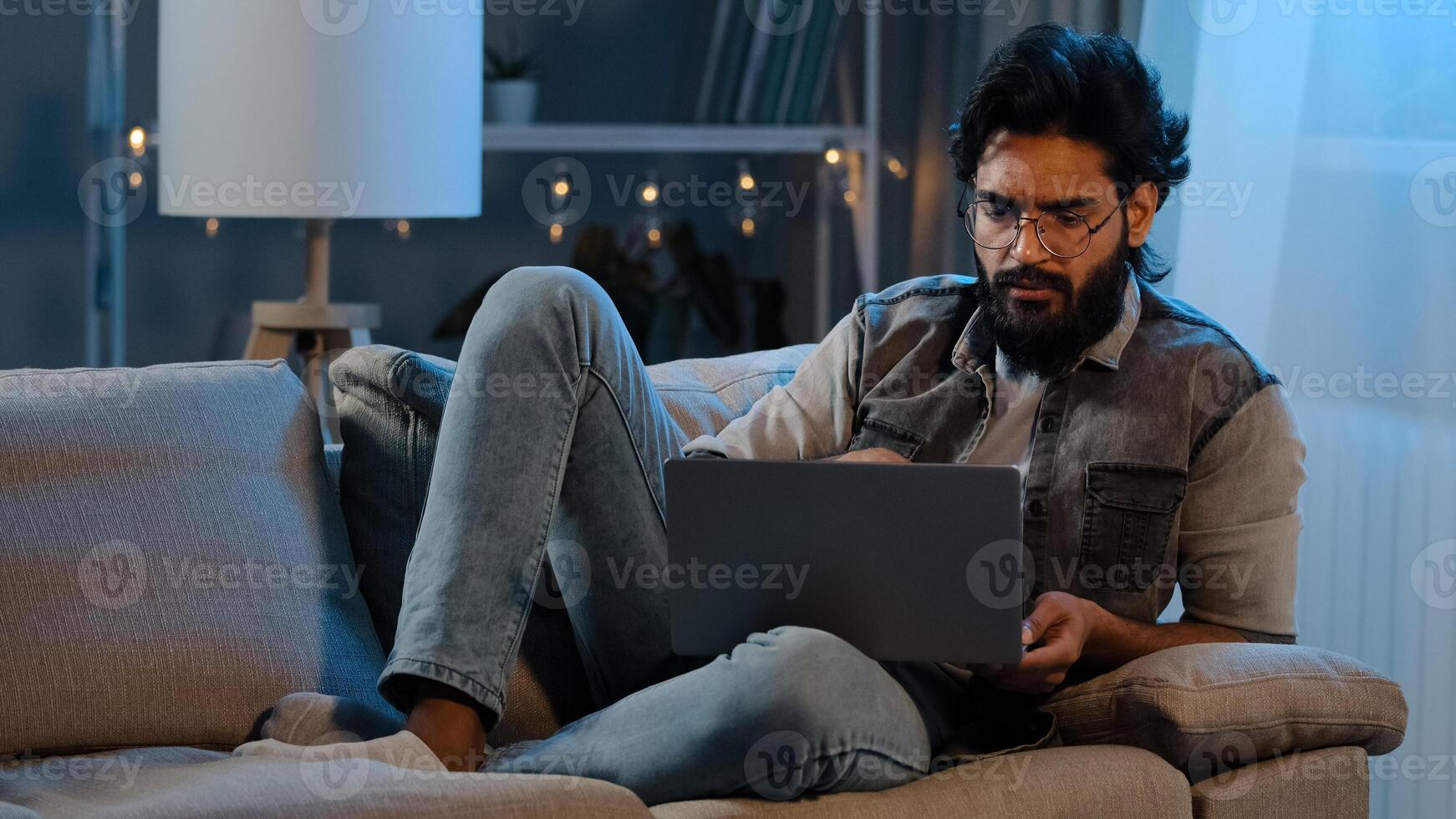 Freelancer guy Arab Indian man bearded male user wearing glasses sitting on sofa at night late time with laptop reading news online thinking doubts job problems pondering solution looking at screen photo
