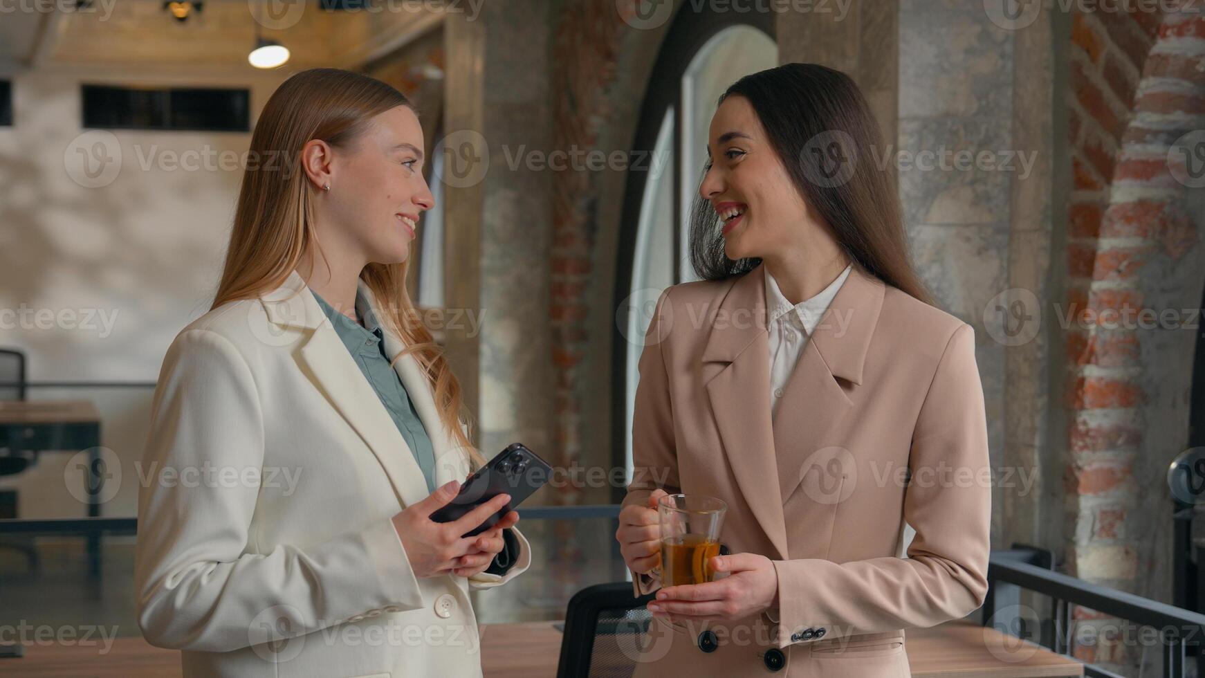 Two happy smiling talking Caucasian women girls office workers female managers businesswomen business friends smile talk conversation work break discuss news hold tea cup mobile phone laugh friendship photo