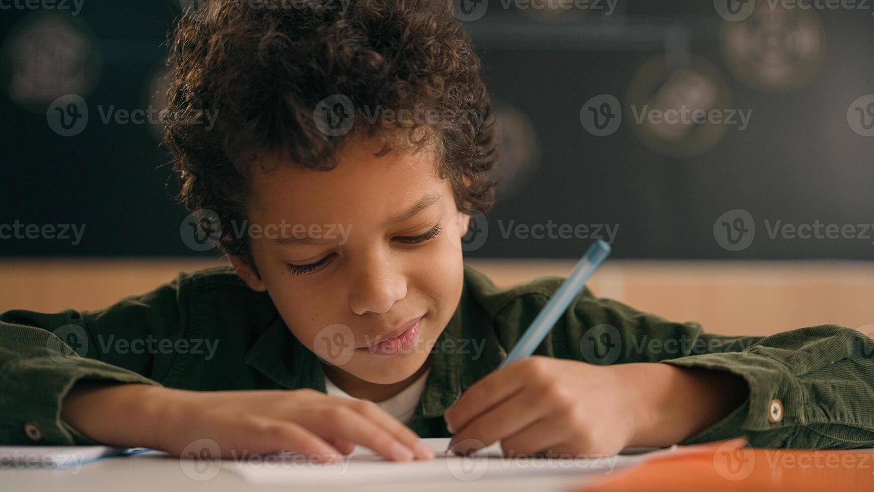 Little African American guy boy writing notes task class lesson primary school write in copybook kid schoolboy son child pupil student learning listen teacher studying education sitting at table desk photo