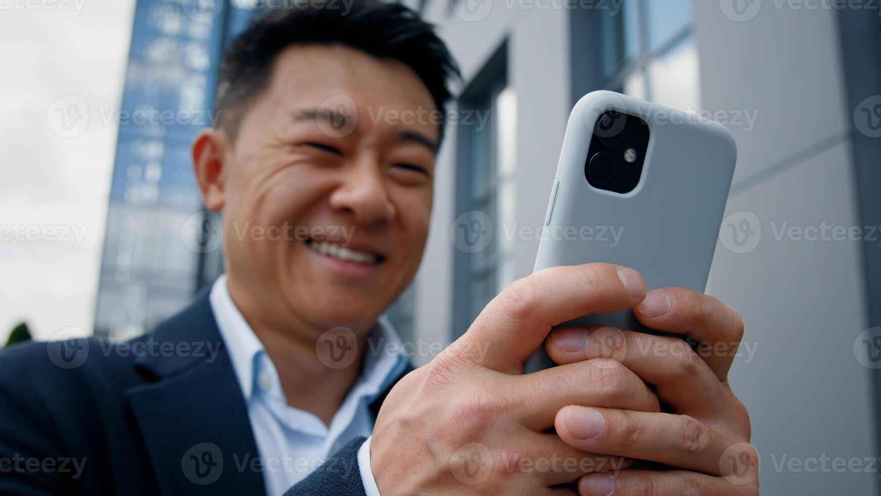 Close up bottom view happy positive smiling formal Asian middle-aged businessman adult 40s worker man employer entrepreneur chatting in mobile phone browsing texting in smartphone outdoors in city photo