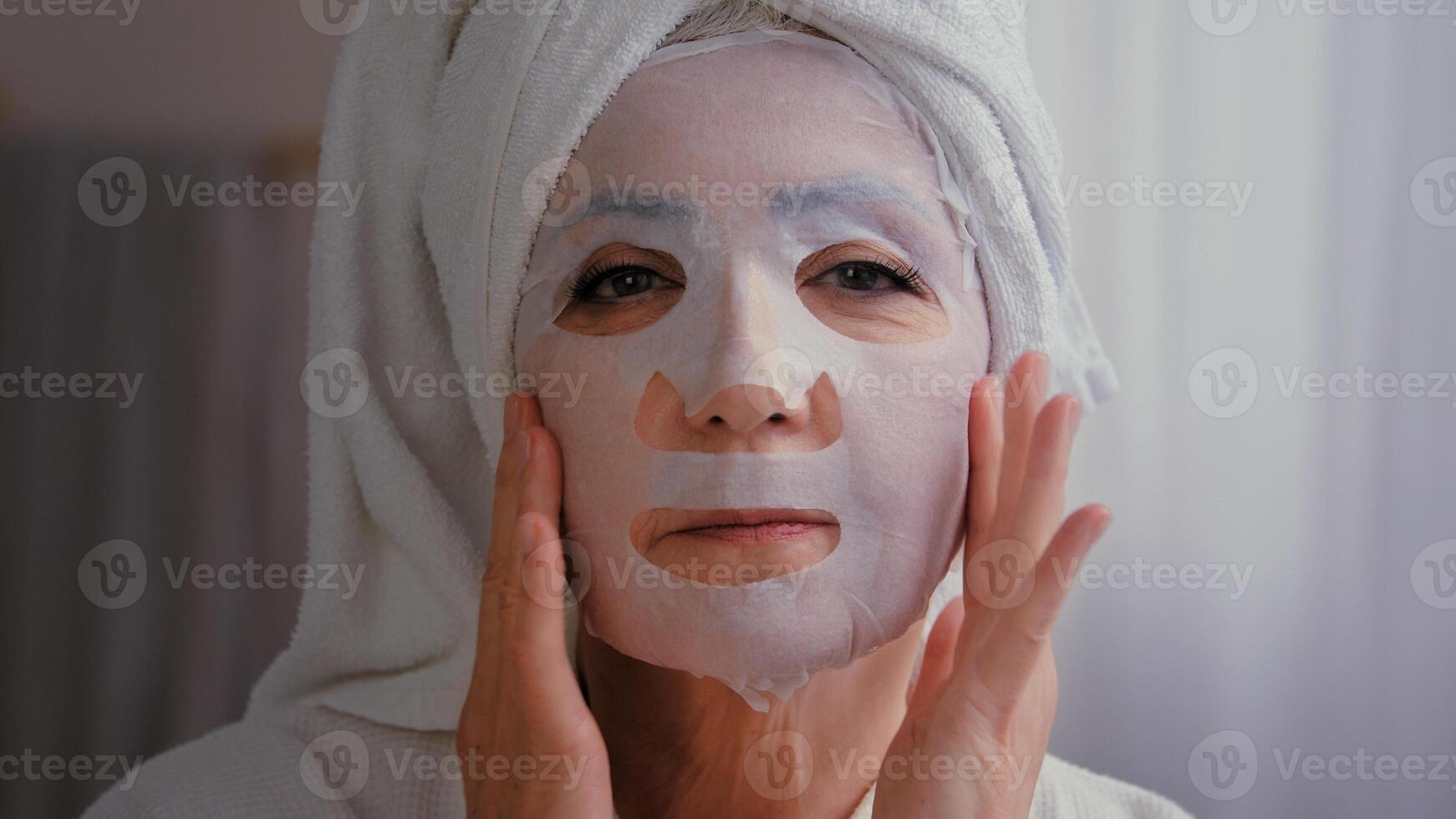 Portrait in bath old senior mature Caucasian woman wrap towel on head grandmother lady apply skincare textile skin mask on female face facial care beauty cosmetology procedure moisturizing cosmetic photo