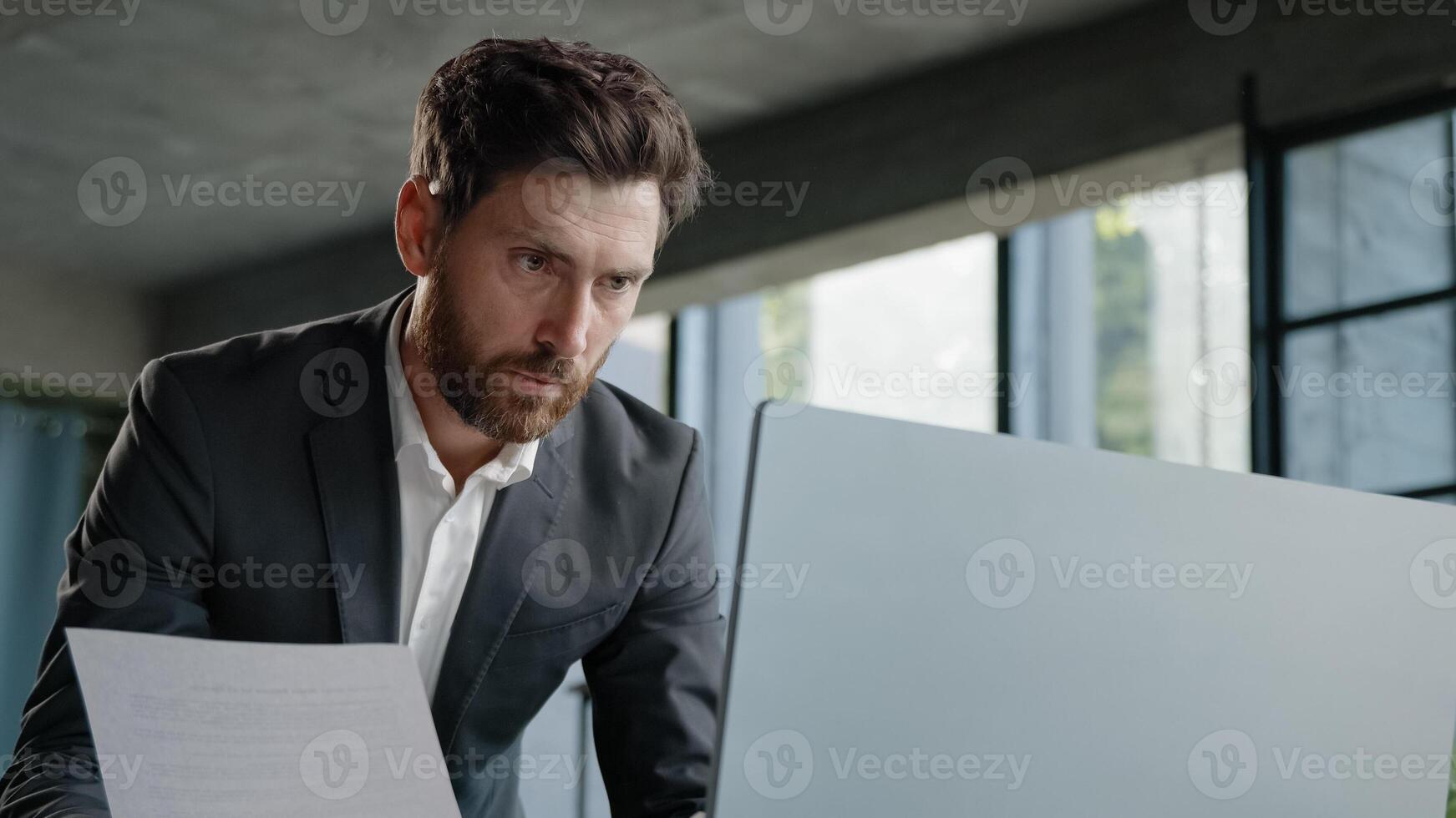 Serious 40s bearded businessman entrepreneur employee standing in office looking at papers report has problem with papers check data in online net service in computer typing working paperwork deadline photo