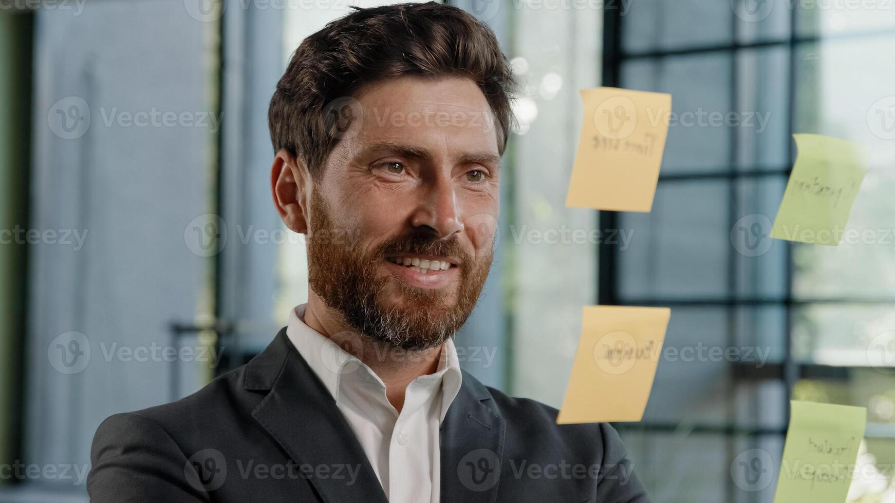 Creative bearded Caucasian 40s businessman boss company CEO leader manager marketing specialist planning management project deals ideas strategy glue sticky notes on transparent glass wall in office photo