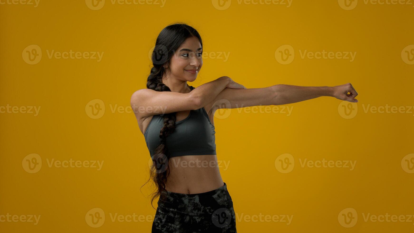 Multiracial woman Indian sporty girl sports trainer sport athlete female in yellow studio talk looking to camera explain exercise showing hand arm and neck head stretching pose stretch workout online photo