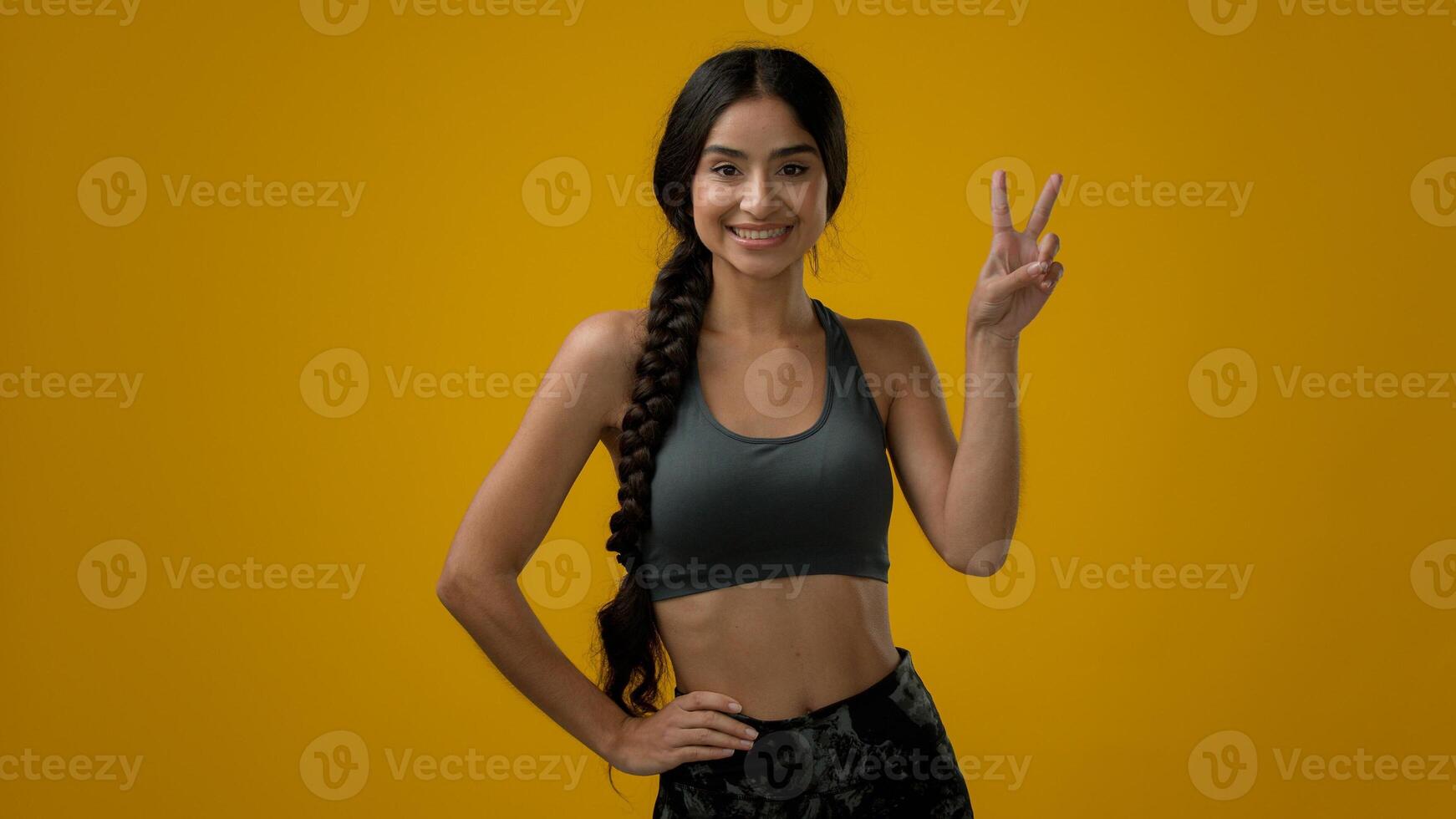 Smiling ethnic Indian sport woman girl lady female athlete in yellow studio background show double fingers gesture two 2 number victory sign peace hand symbol showing V gesturing arm win twice second photo