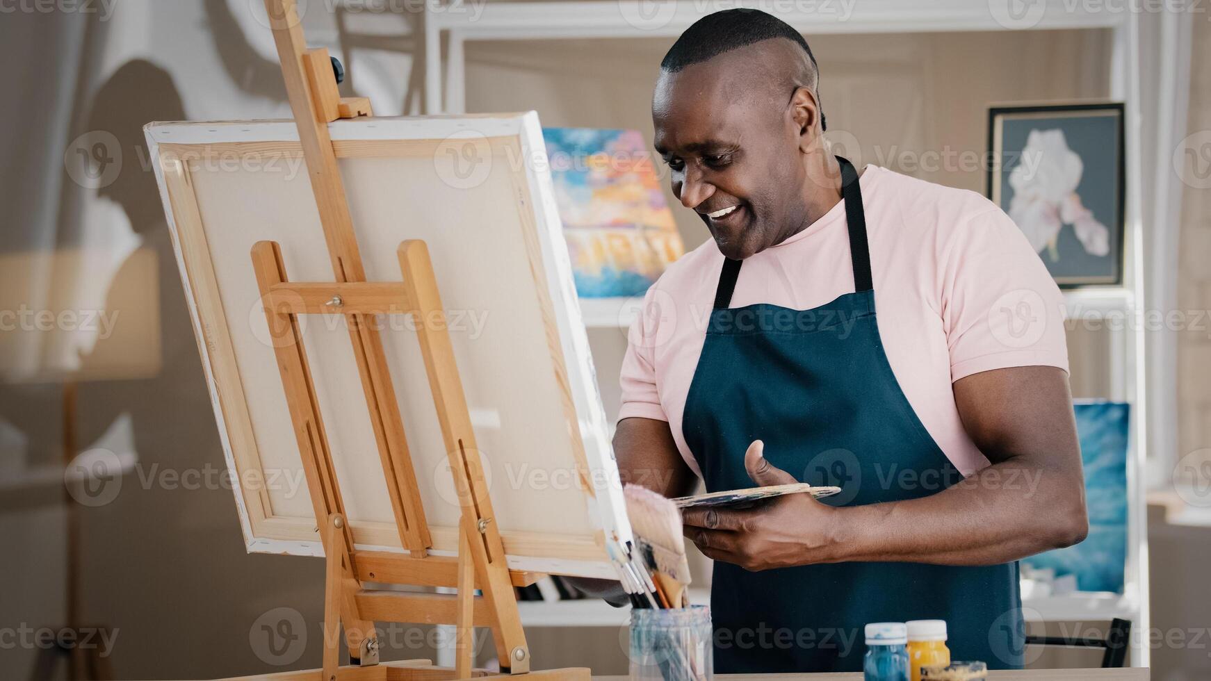 Side view happy smiling cheerful creative talented inspired old African American senior man artist male painter in apron drawing painting picture creative idea on canvas in art studio school paints photo