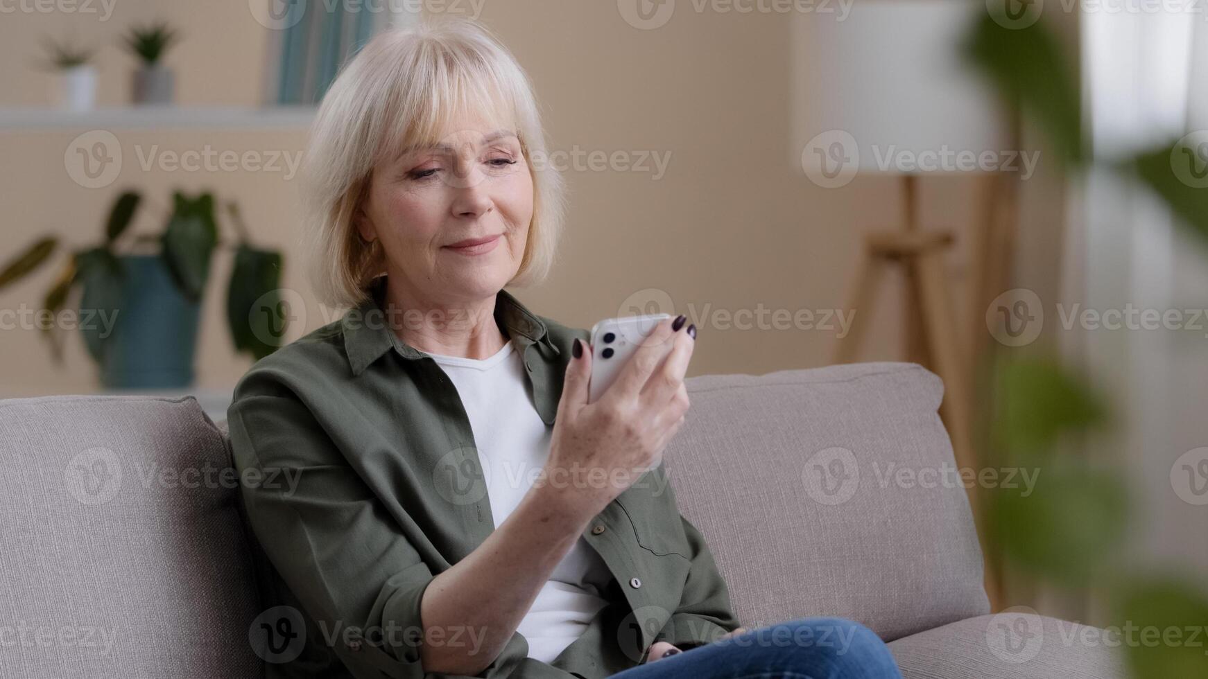 Old Caucasian woman 60s elderly mature lady sits at home on sofa with mobile phone reads news receives notice thinks dreams ponders idea looking away deep in thoughts thinking problem with modern tech photo