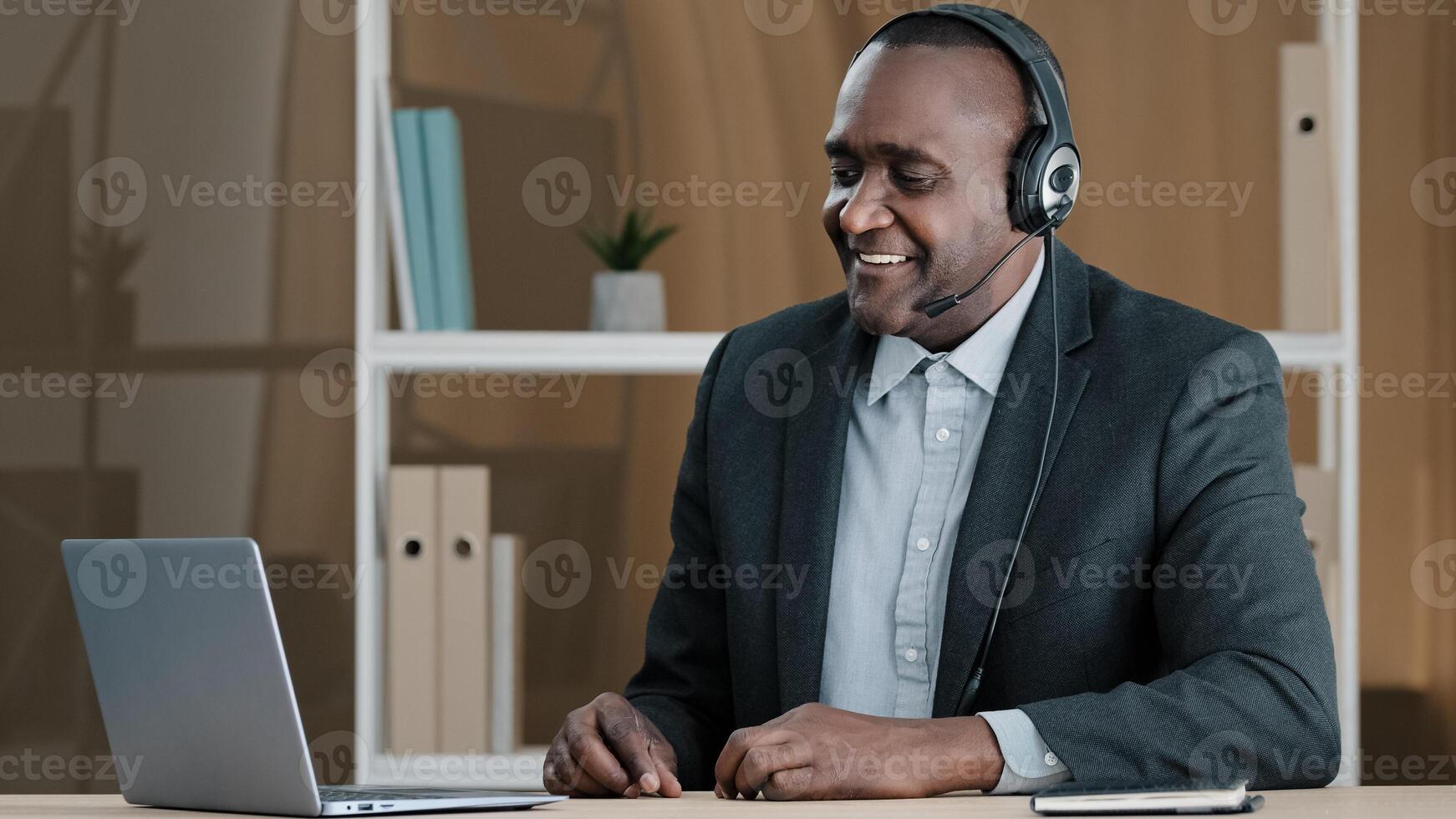 Elderly cheerful businessman African agent broker salesman old 50s man boss in headset has virtual meeting with client talking remote from office call conference using laptop app laughing happy photo