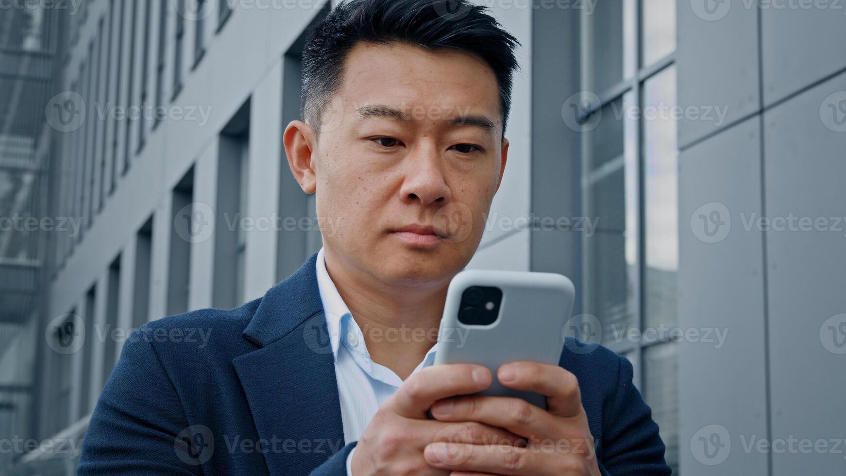 Bottom view pensive Asian Korean adult man middle-aged 40s businessman standing in city near office building looking up thinking browsing typing chatting in smartphone mobile phone web ordering online photo