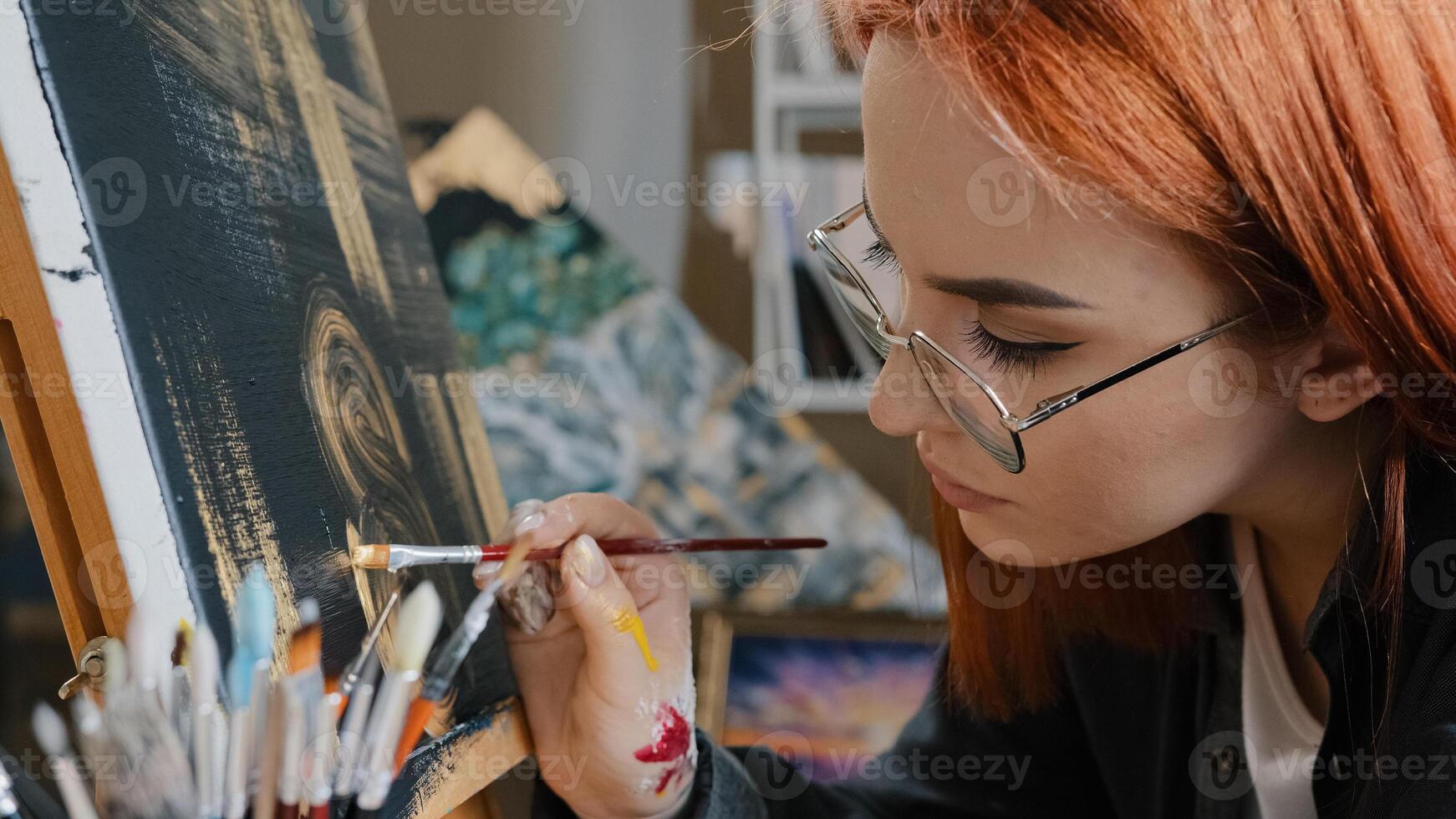 Side view red-haired painter artist creator designer woman student girl in glasses holding brush paints picture on black background with golden oil learning art painting on canvas yellow abstraction photo