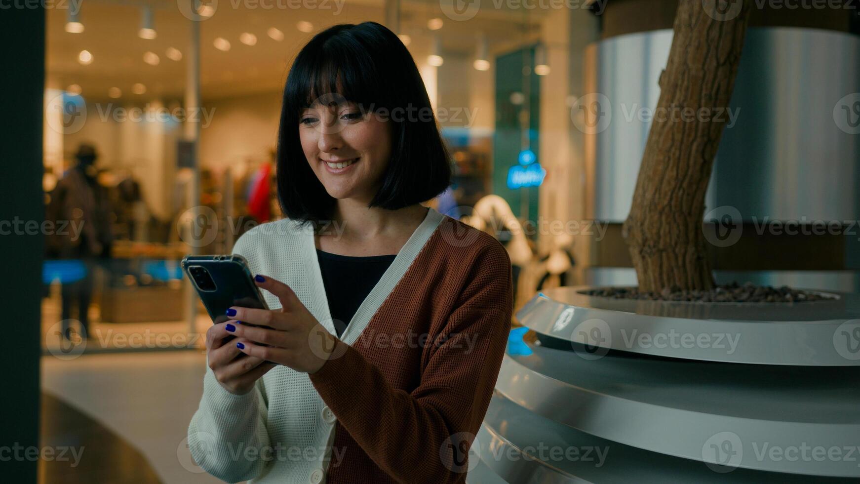 Smiling 20s woman lady female girl indoors in shopping mall sitting looking at mobile phone screen scrolling browsing in net app with smartphone smile chatting in social media e-commerce buying online photo