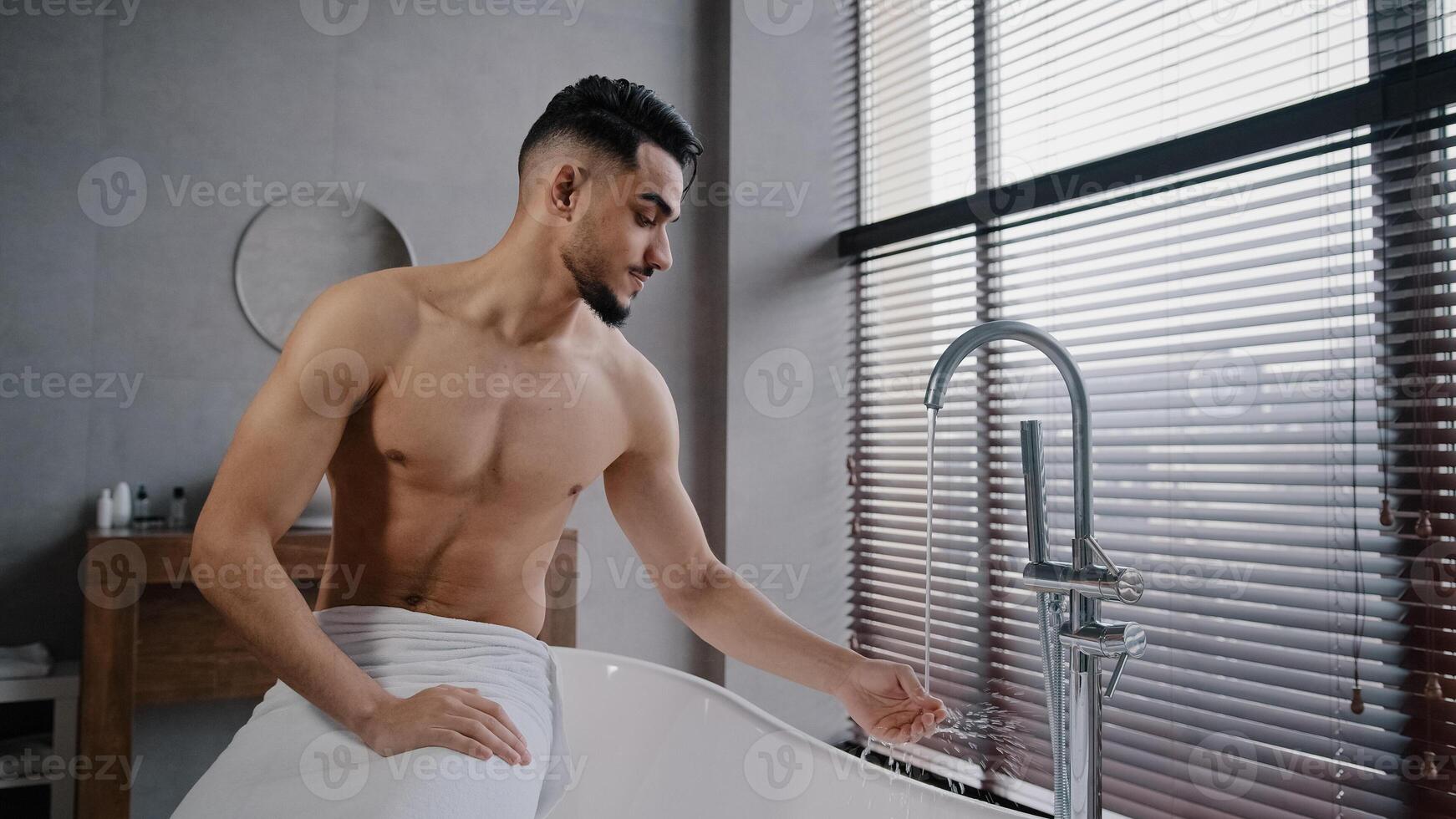 Millennial bare naked muscular indian arab man guy with beard wrapped towel around hips sitting on bathrub in bathroom at home turning faucet tasting hot cold water with hand getting ready for bathing photo