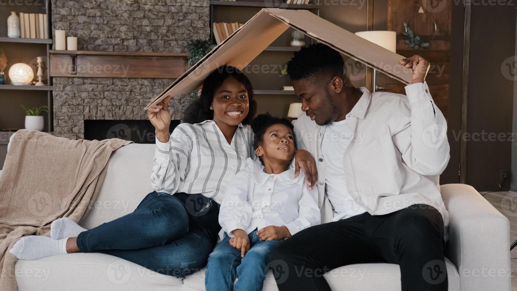 African american family parents with daughter little girl adopted child sit on sofa under cardboard roof dreaming about own house relocation home insurance real estate buying flat kid protection photo