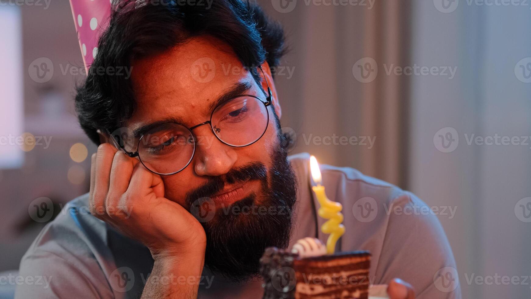 Headshot arabian alone single sad upset lonely indian bearded man in festive birthday pink hat and glasses sitting at home at night feel frustrated despair disappointed sadness holds cake with candle photo